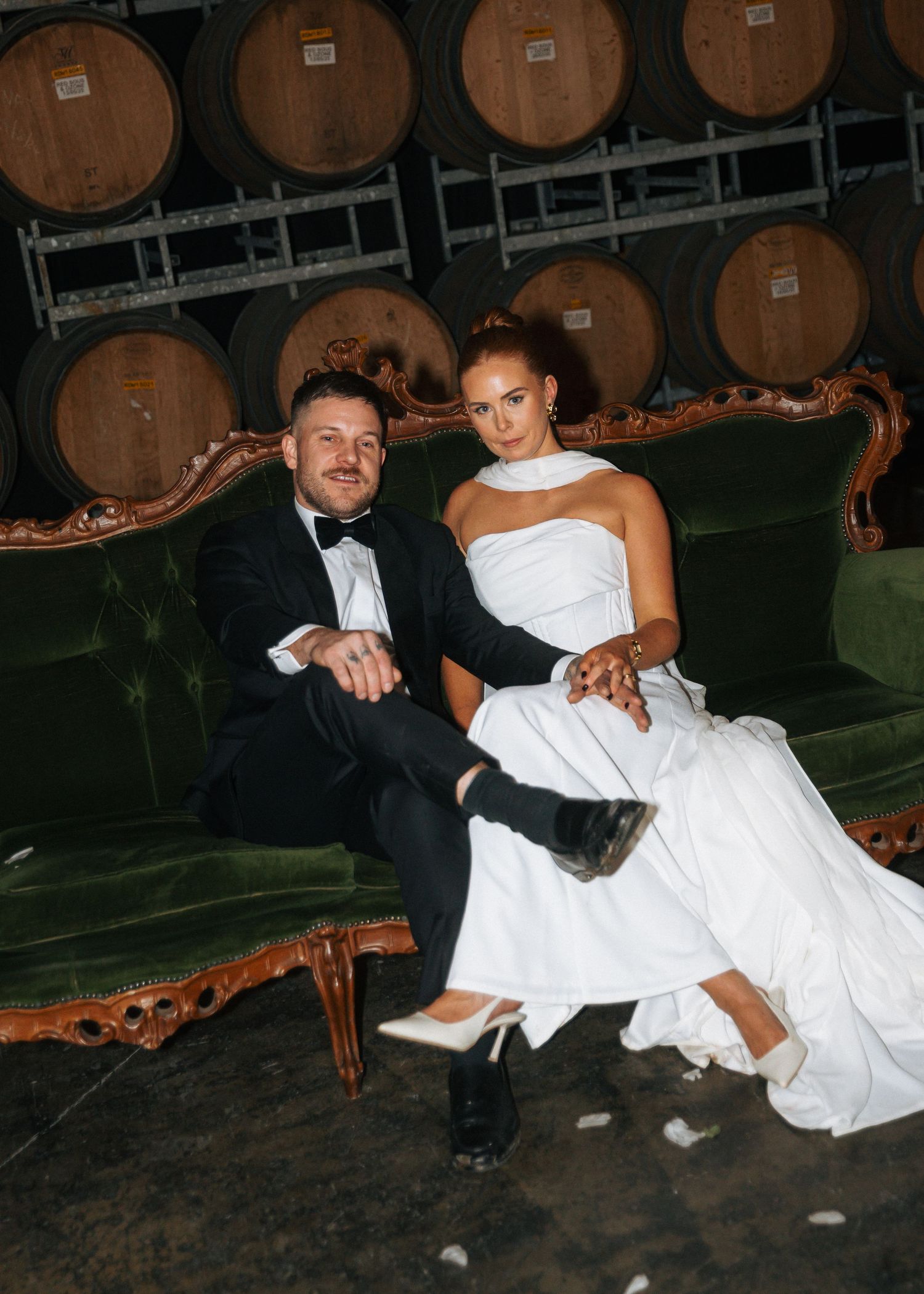 Bride and groom relaxing on vintage green velvet sofa surrounded by wine barrels at winery.