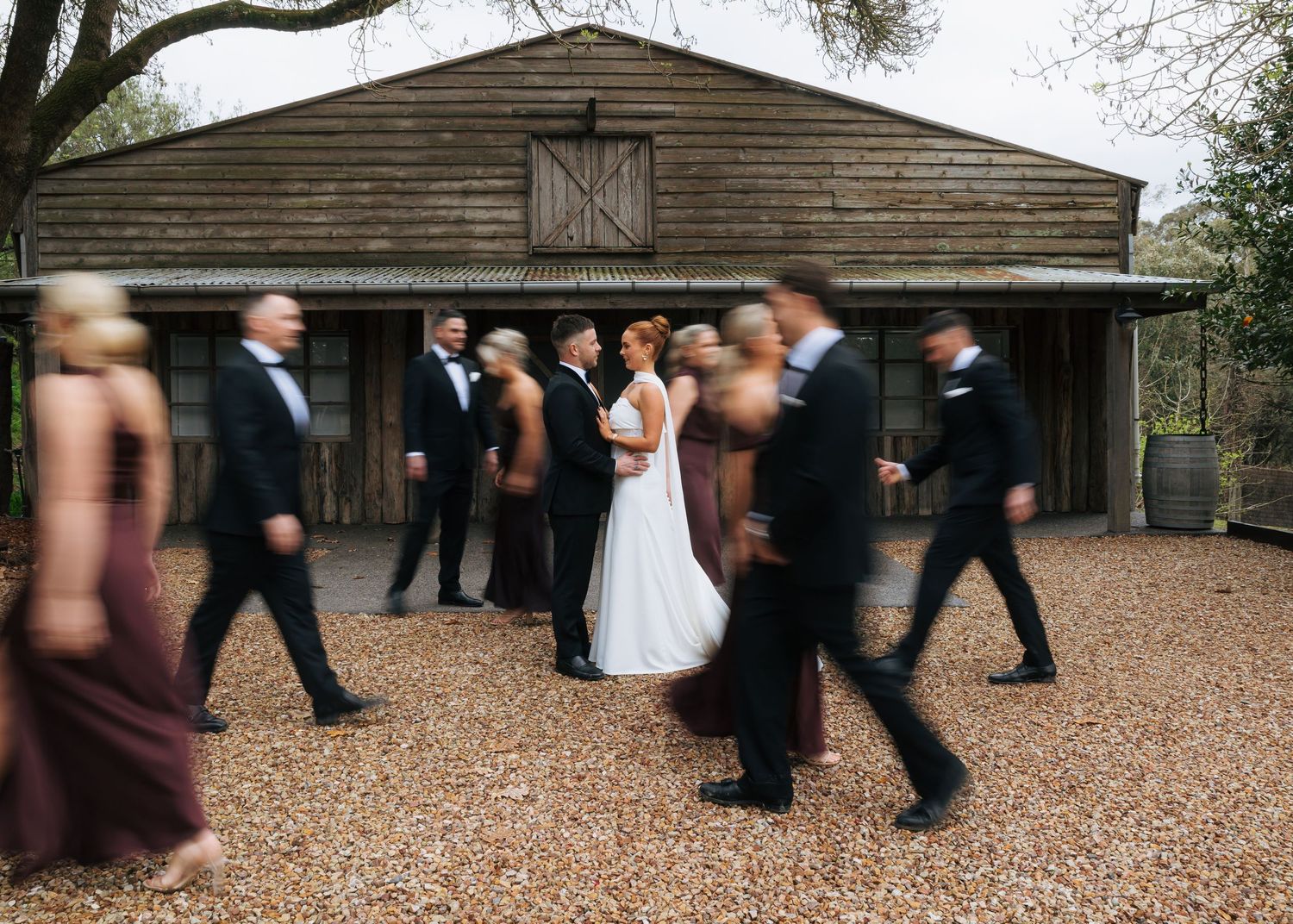 Wedding party walking past rustic barn venue, bride and groom standing still amid motion-blurred bridesmaids and groomsmen.