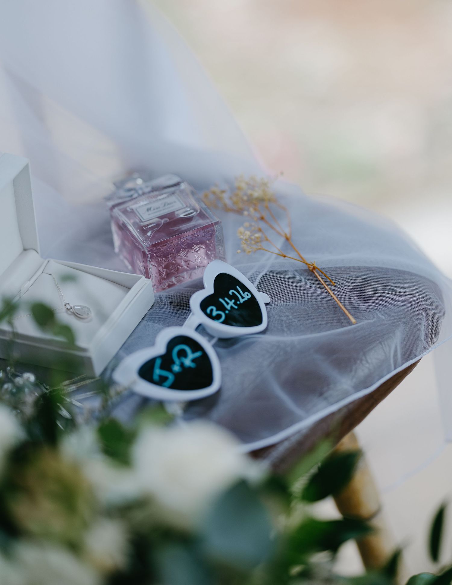 Wedding flat lay with heart-shaped sunglasses, pink perfume bottle, ring box, and veil on a chair.