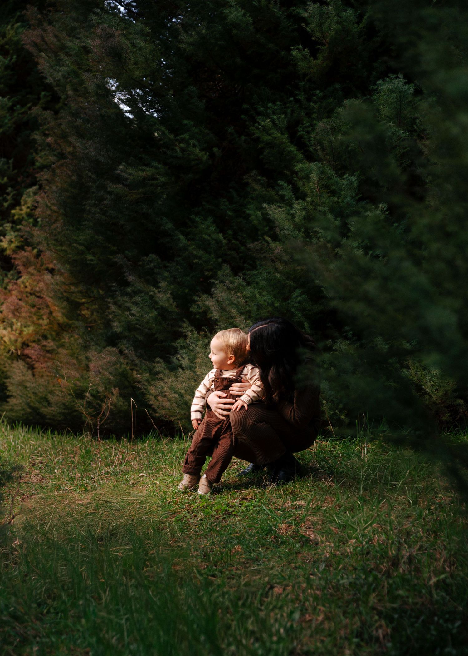 Pregnant mother crouching to embrace young child in dappled forest light.