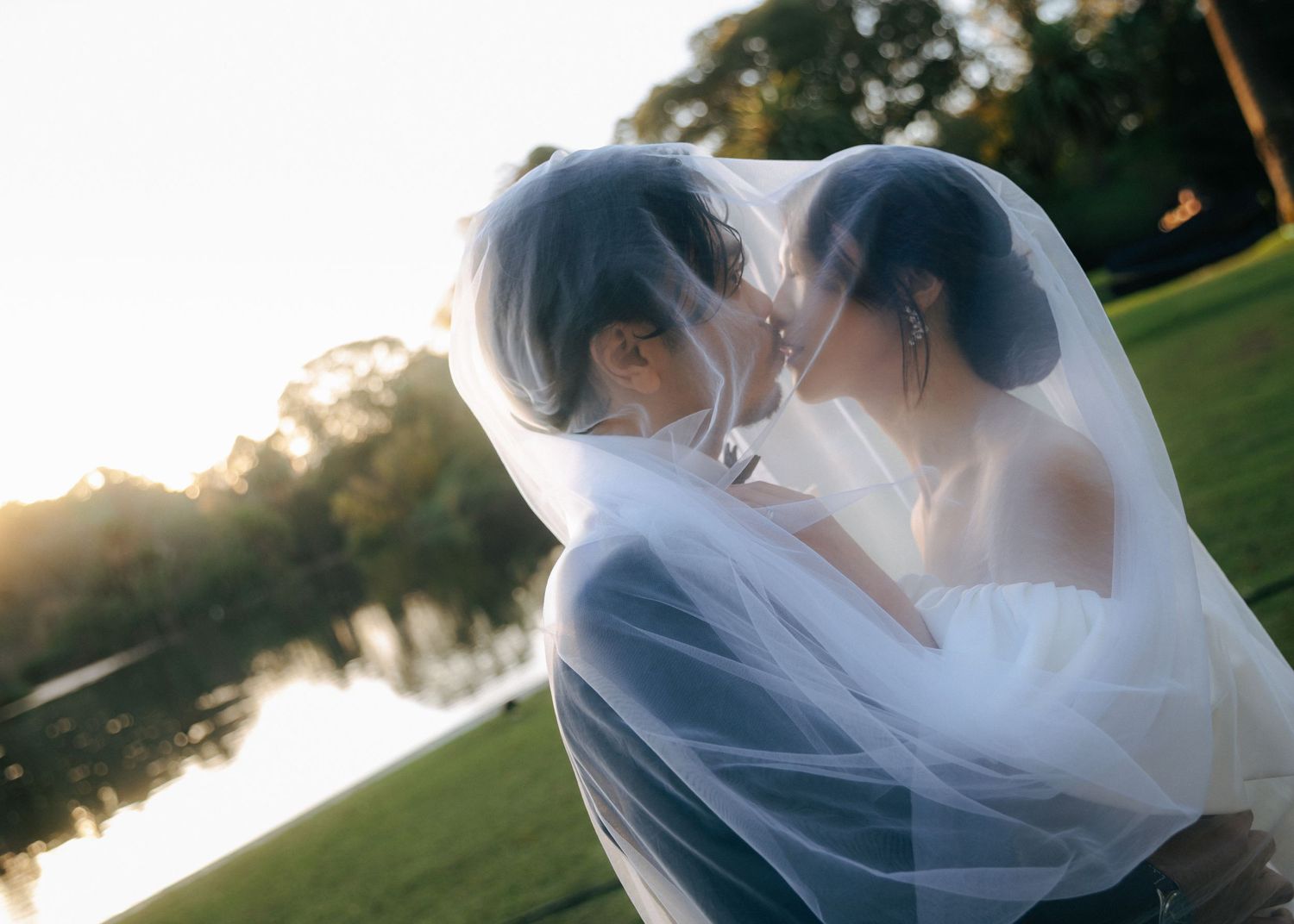 A bride and groom share a romantic kiss under a flowing veil at golden hour by a serene lake.