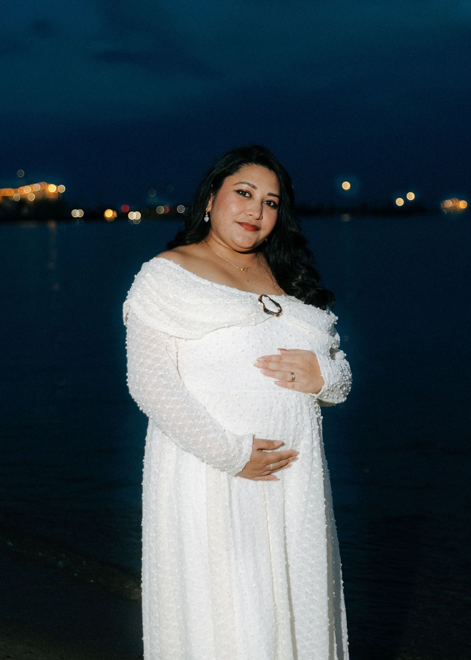 Pregnant woman in white textured off-shoulder dress cradling belly near water at night.