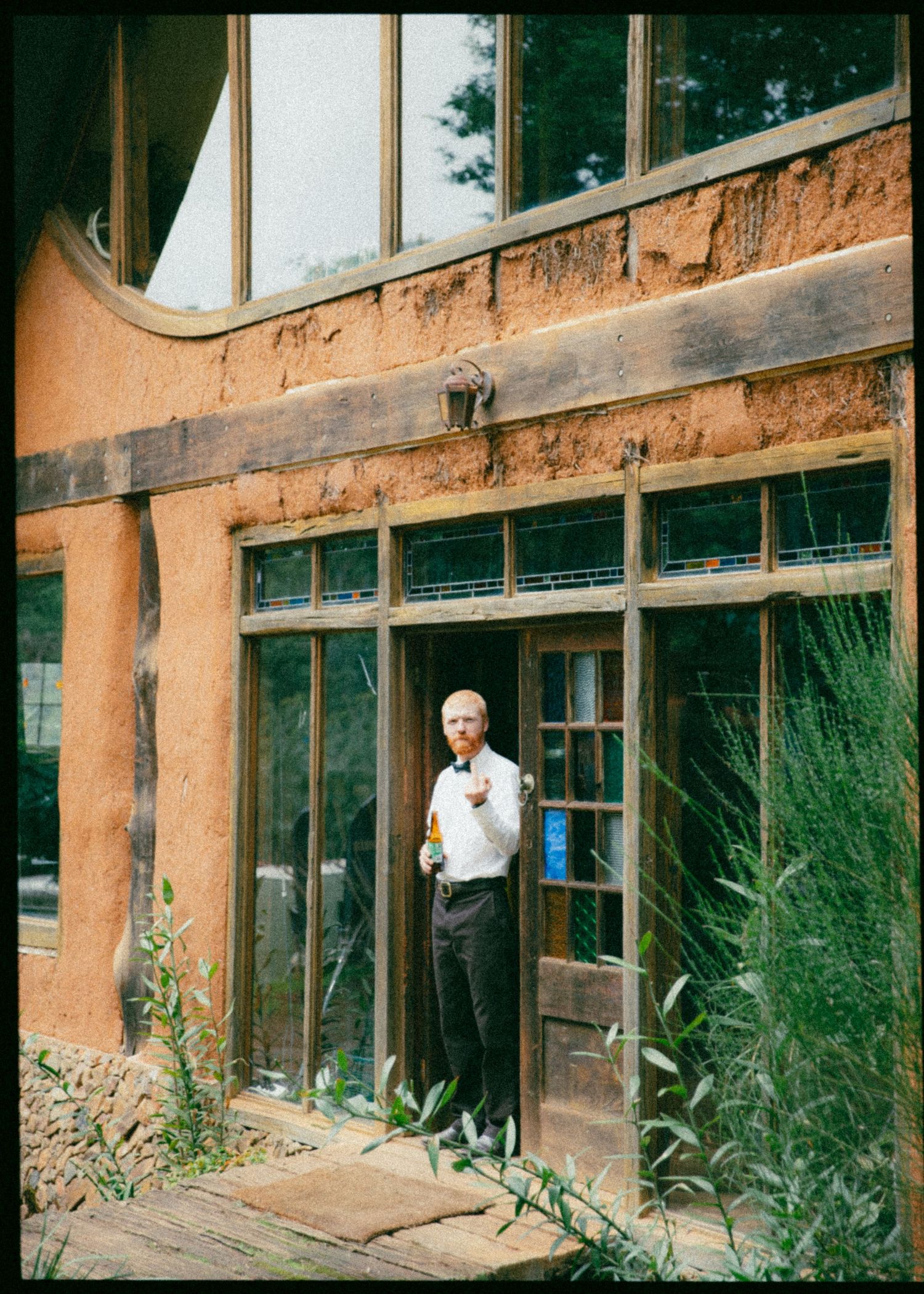 Elderly man in white shirt stands outside a unique adobe-style building with large glass windows.