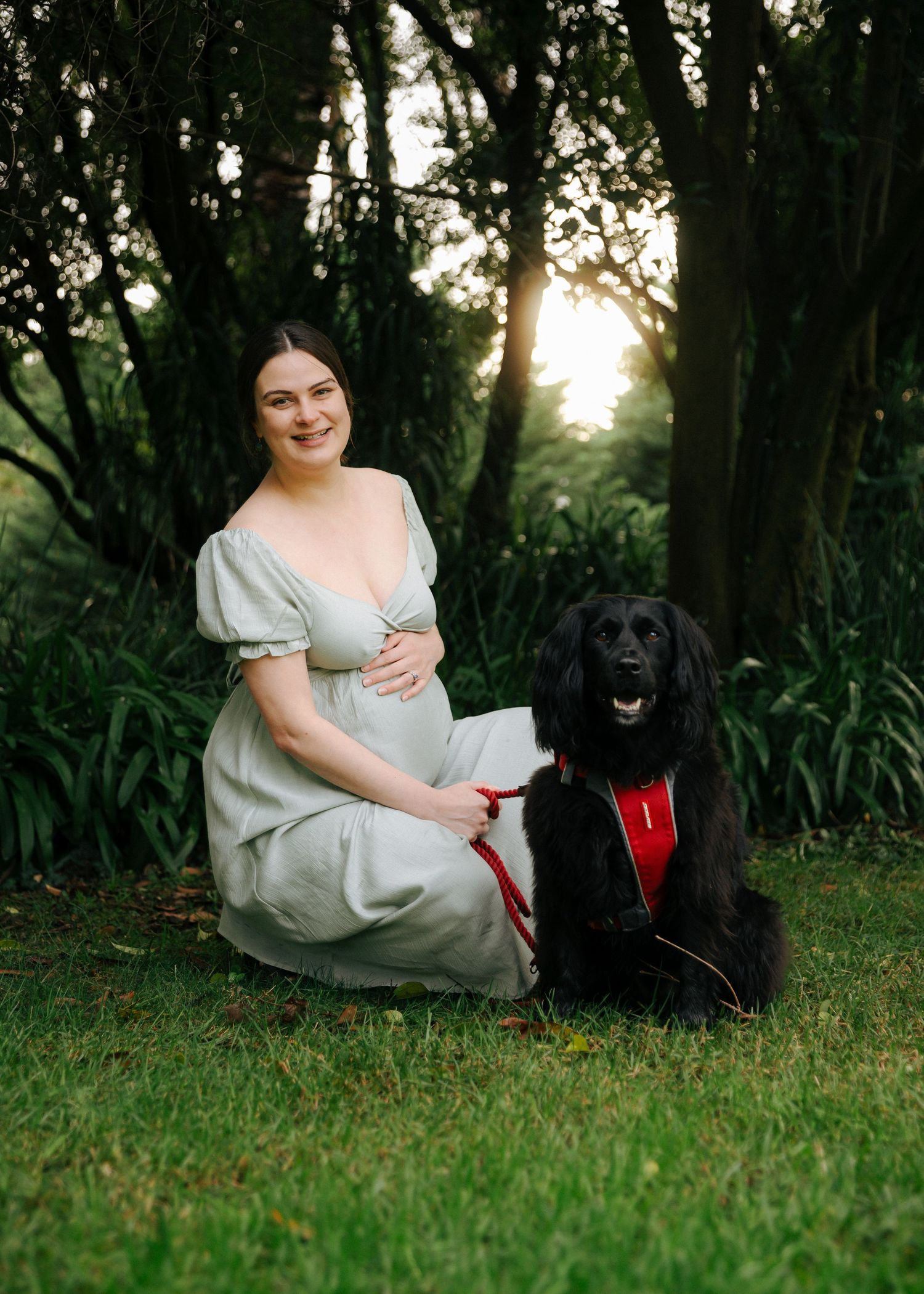Pregnant woman in sage green dress sitting on grass with black dog at golden hour.