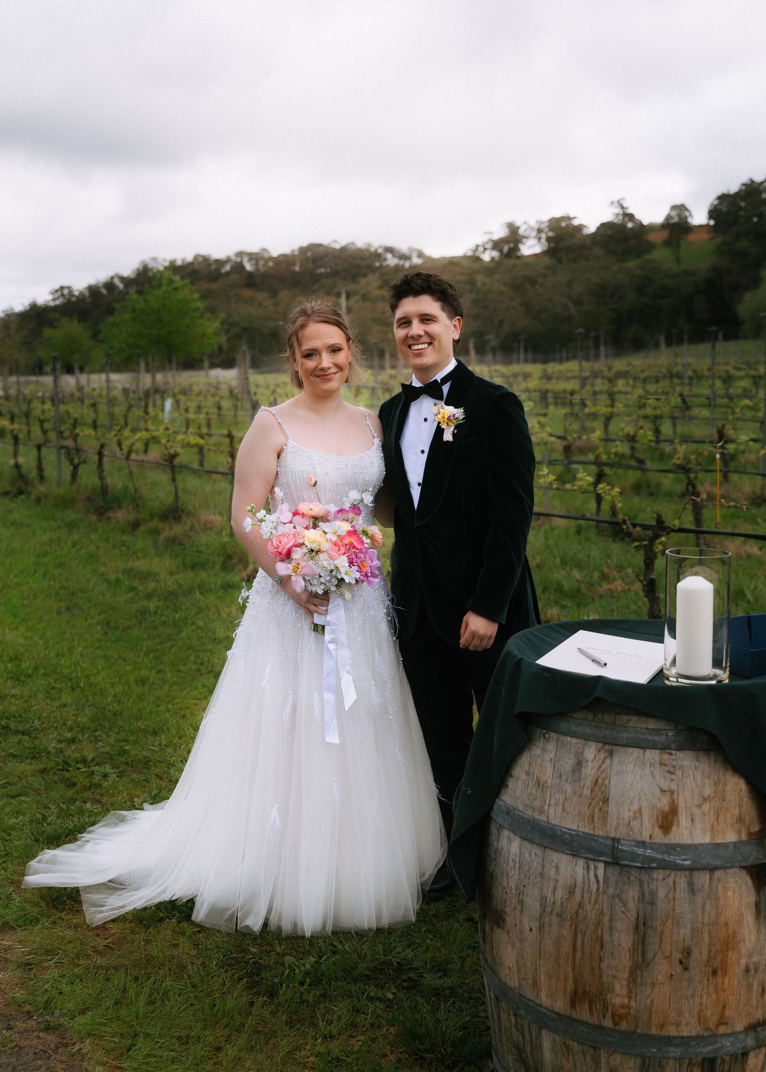 Bride in a tulle gown with a pastel bouquet and groom in a black tuxedo pose in a vineyard beside a barrel with a candle.