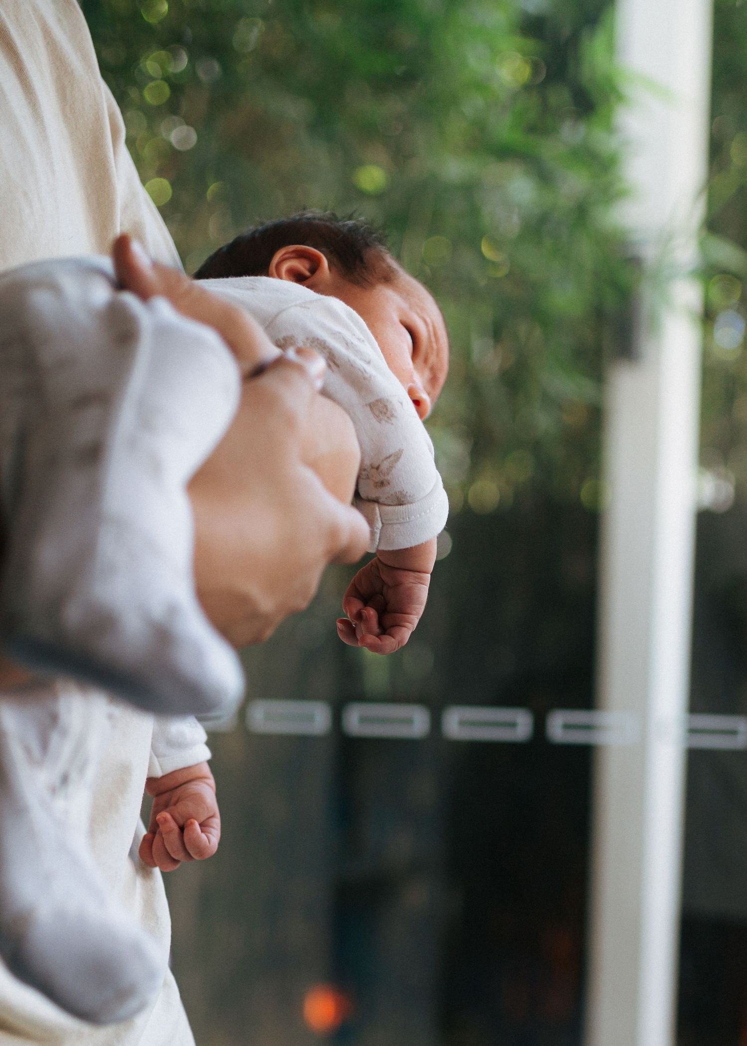 A sleeping newborn baby in a white outfit is cradled in an adult's arms near a glass door with soft green foliage in the background.