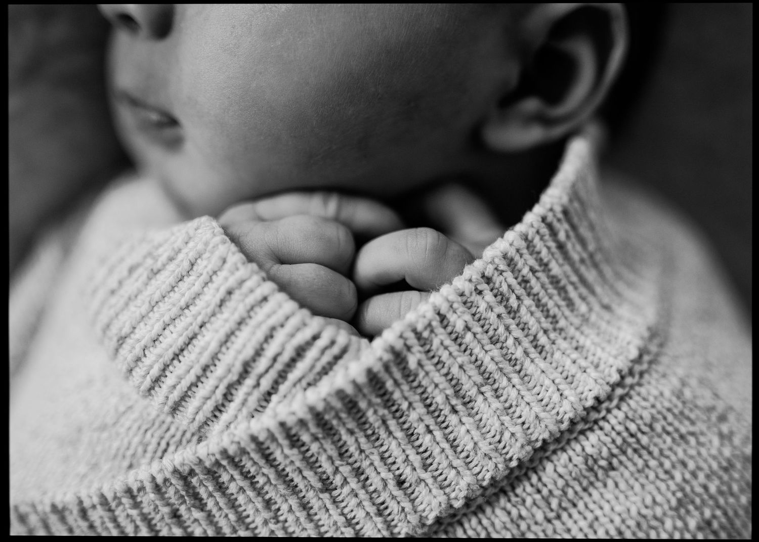 Black and white close-up of a newborn baby's tiny hands tucked under the chin, wrapped in a cozy ribbed knit sweater.