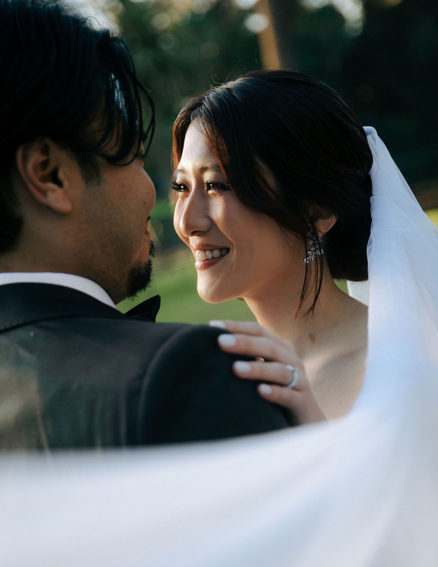 Smiling Asian bride in veil gazes at groom in black tuxedo during golden hour.