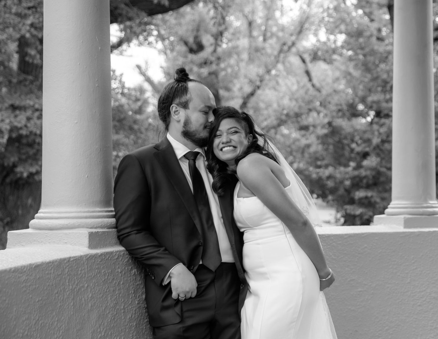 Black and white photo of bride and groom smiling beside columns in a pavilion.