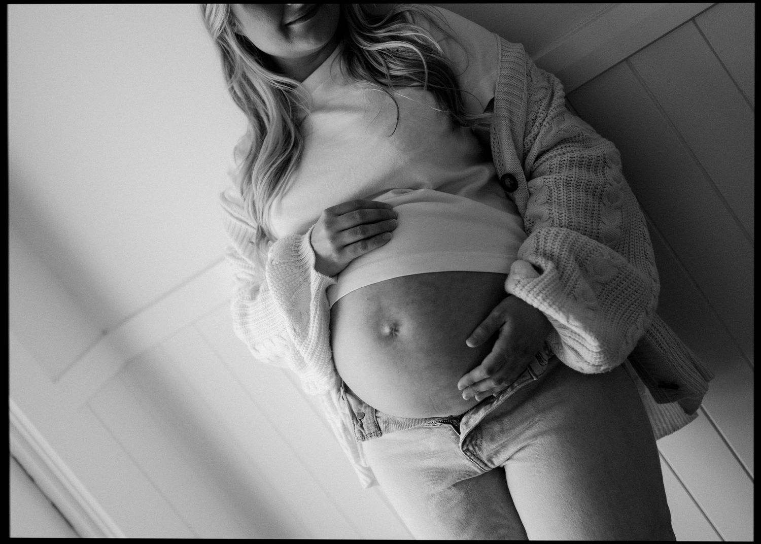 Black and white photo of pregnant woman holding baby bump wearing open cardigan and jeans.