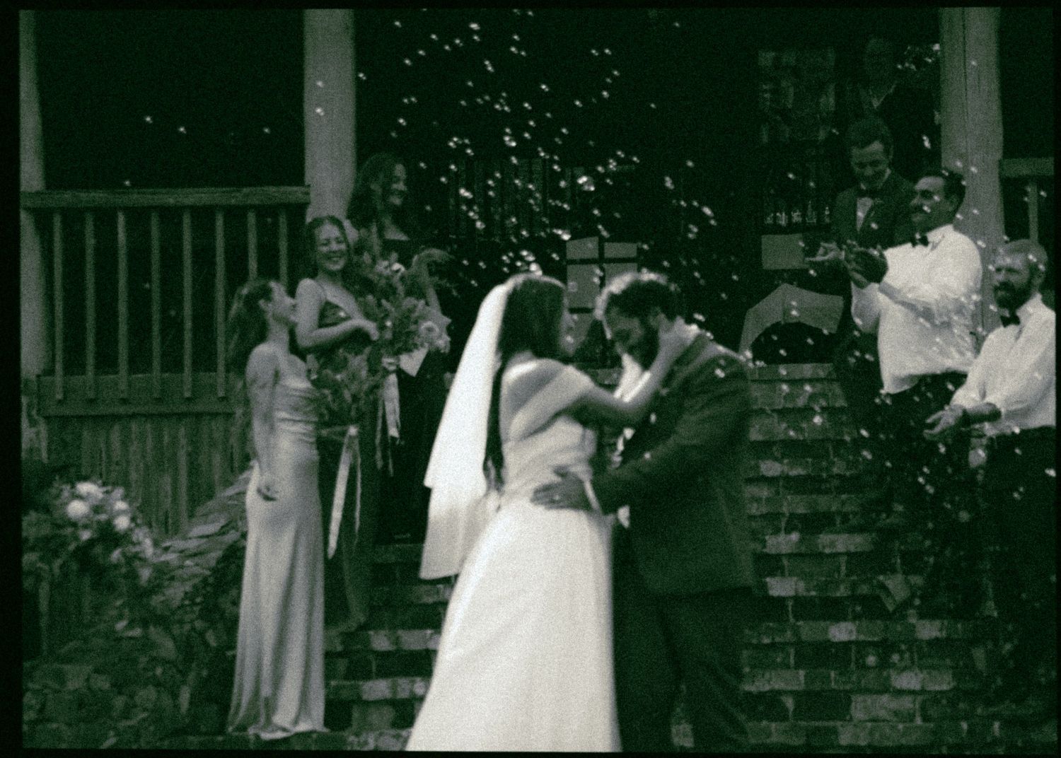 Black and white photo of bride and groom kissing on steps as guests throw confetti.