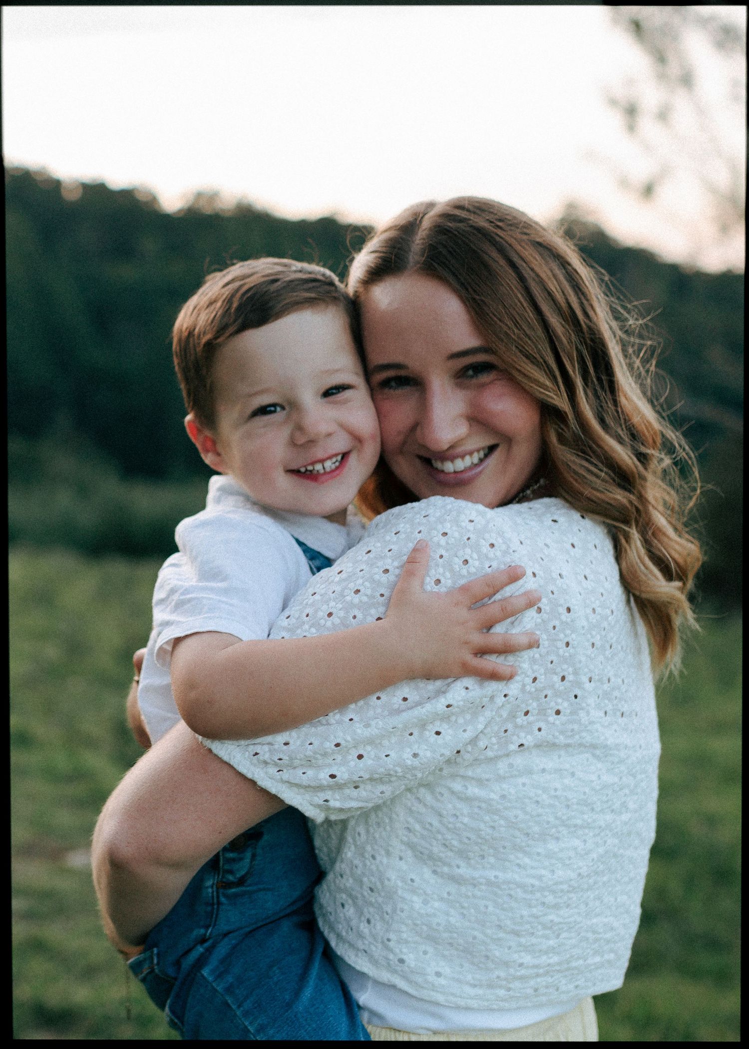 Joyful mother embracing laughing young son outdoors in a lush green meadow at sunset.