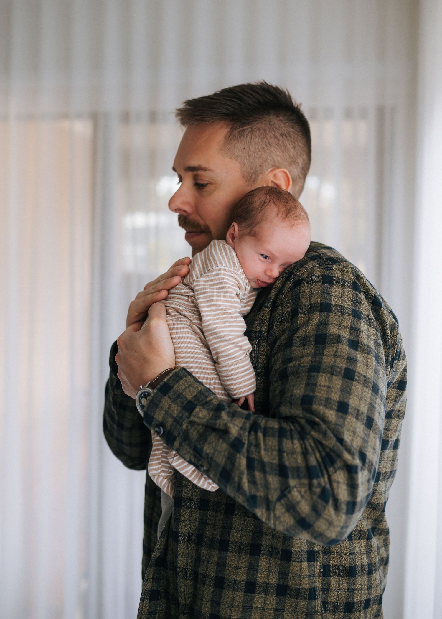 A father in a plaid shirt gently holds his sleeping newborn baby against his shoulder.