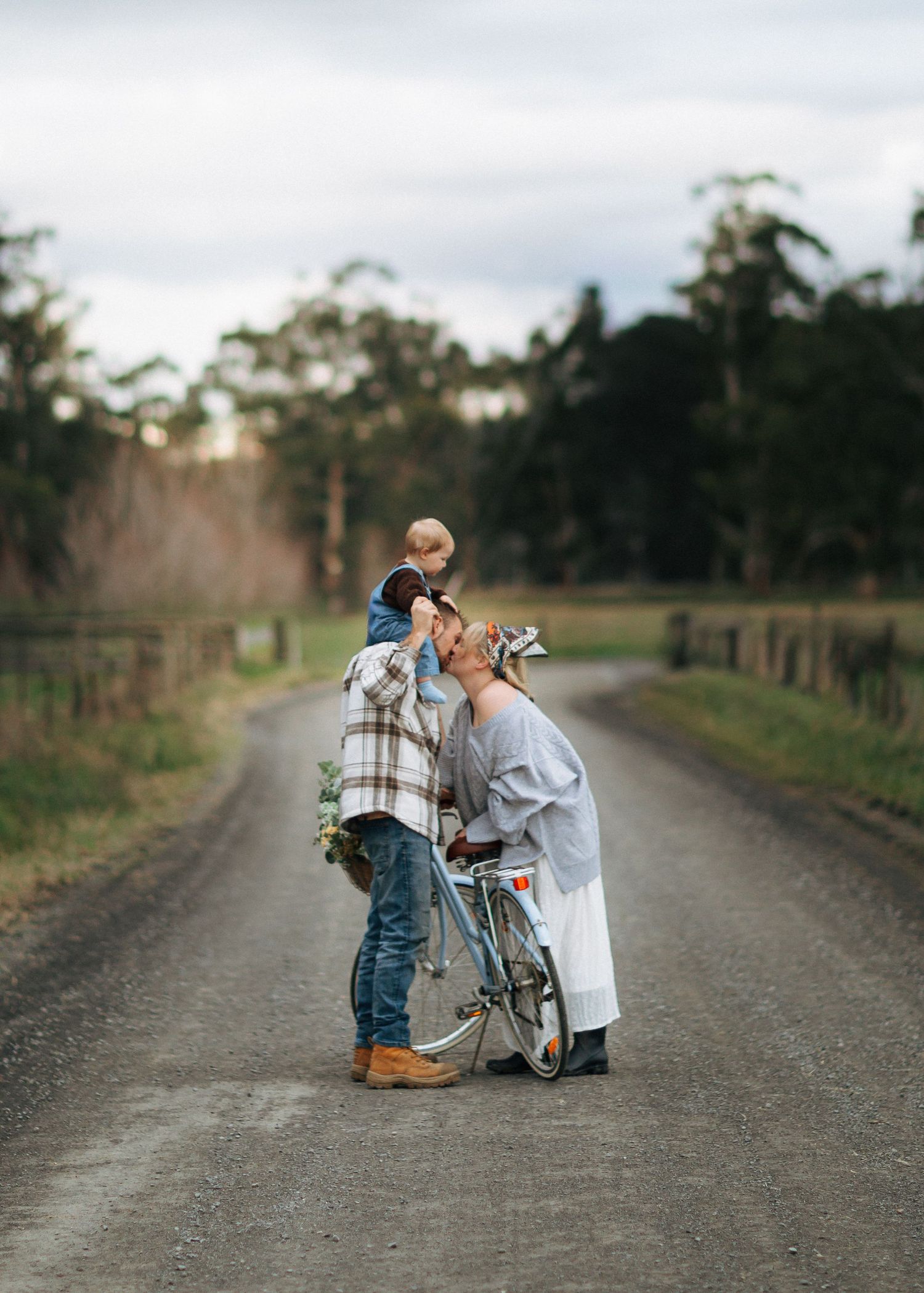 Parents sharing a tender kiss with toddler on fathers shoulders on a quiet rural country road.