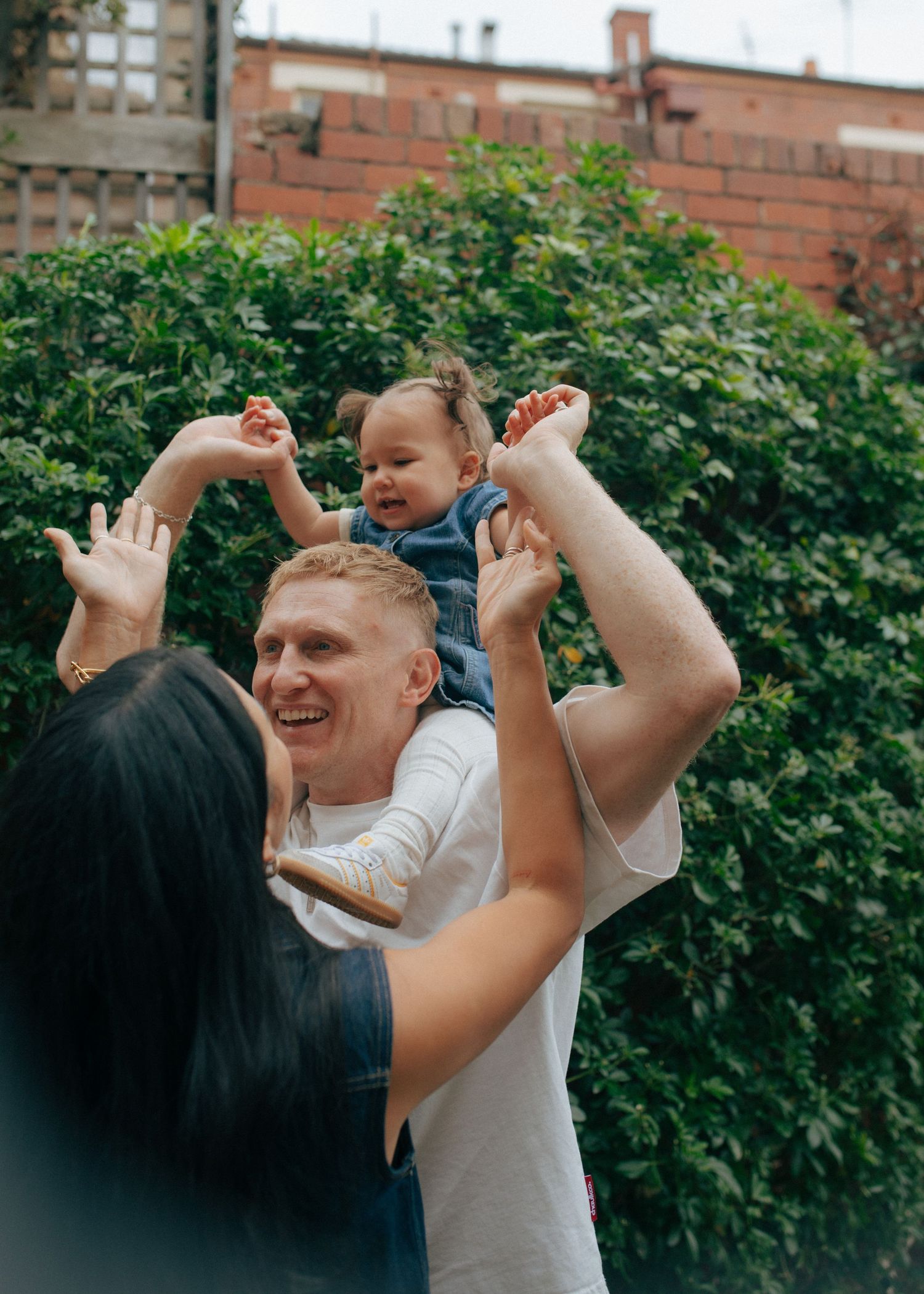 Happy father lifts smiling baby above his head while mother reaches up in a garden.