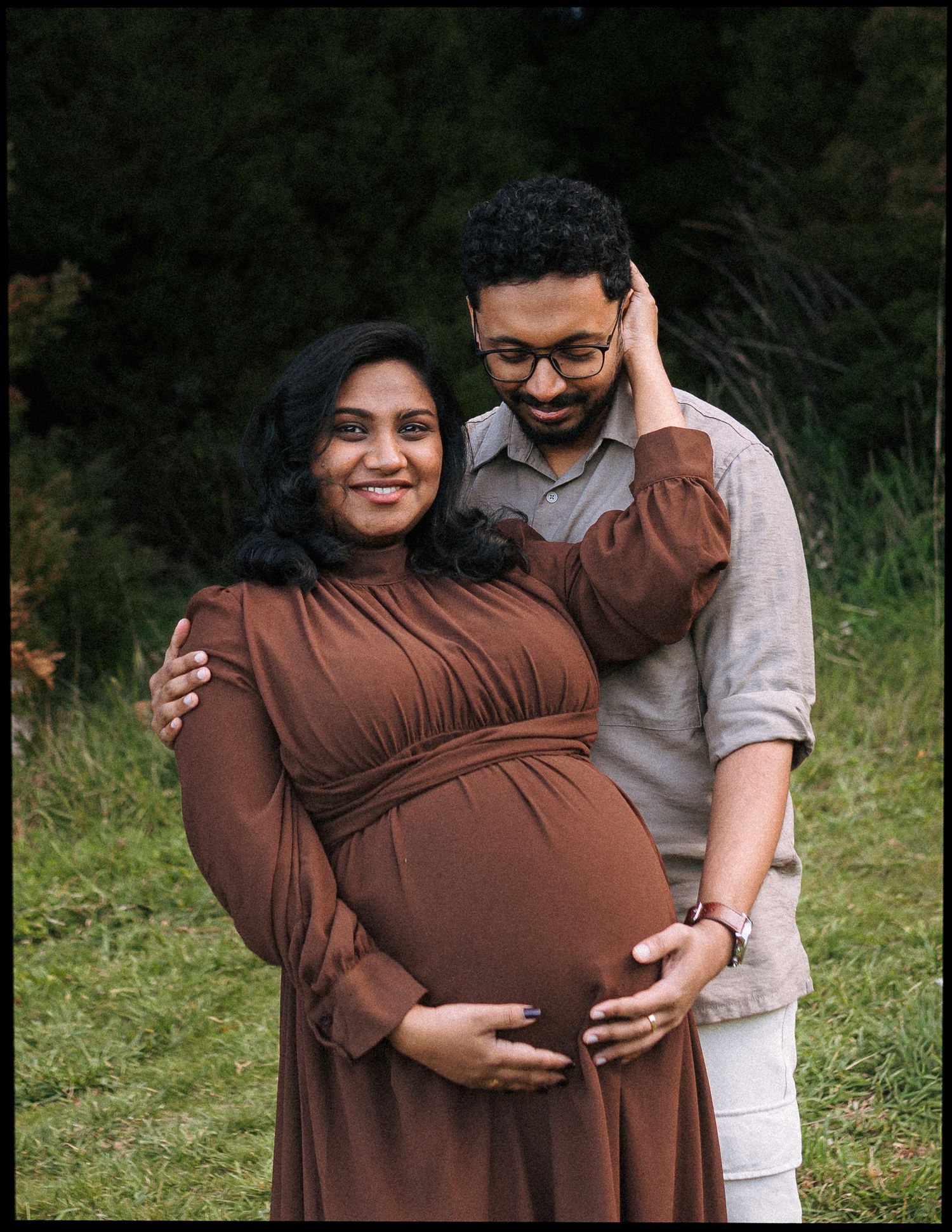 Smiling pregnant woman in a brown dress cradles her baby bump while her bearded husband in glasses embraces her outdoors on a grassy field.