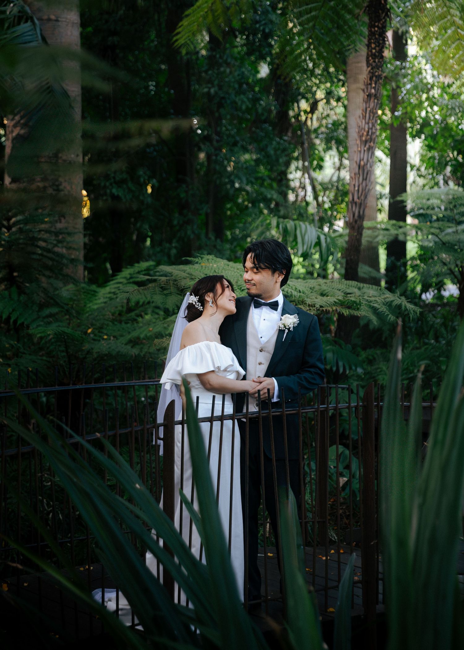 Newlyweds share an intimate moment on a garden bridge, surrounded by tropical ferns and greenery.