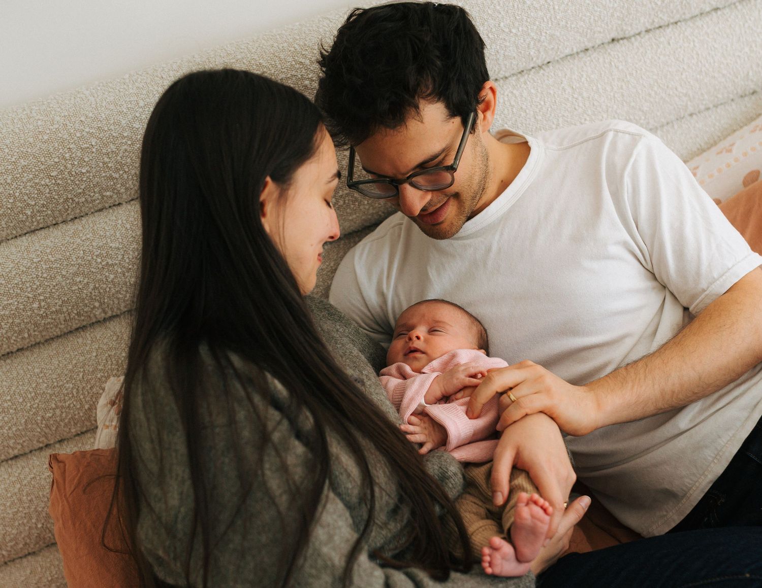 Smiling parents in casual clothes sitting together admiring their newborn baby in pink outfit.