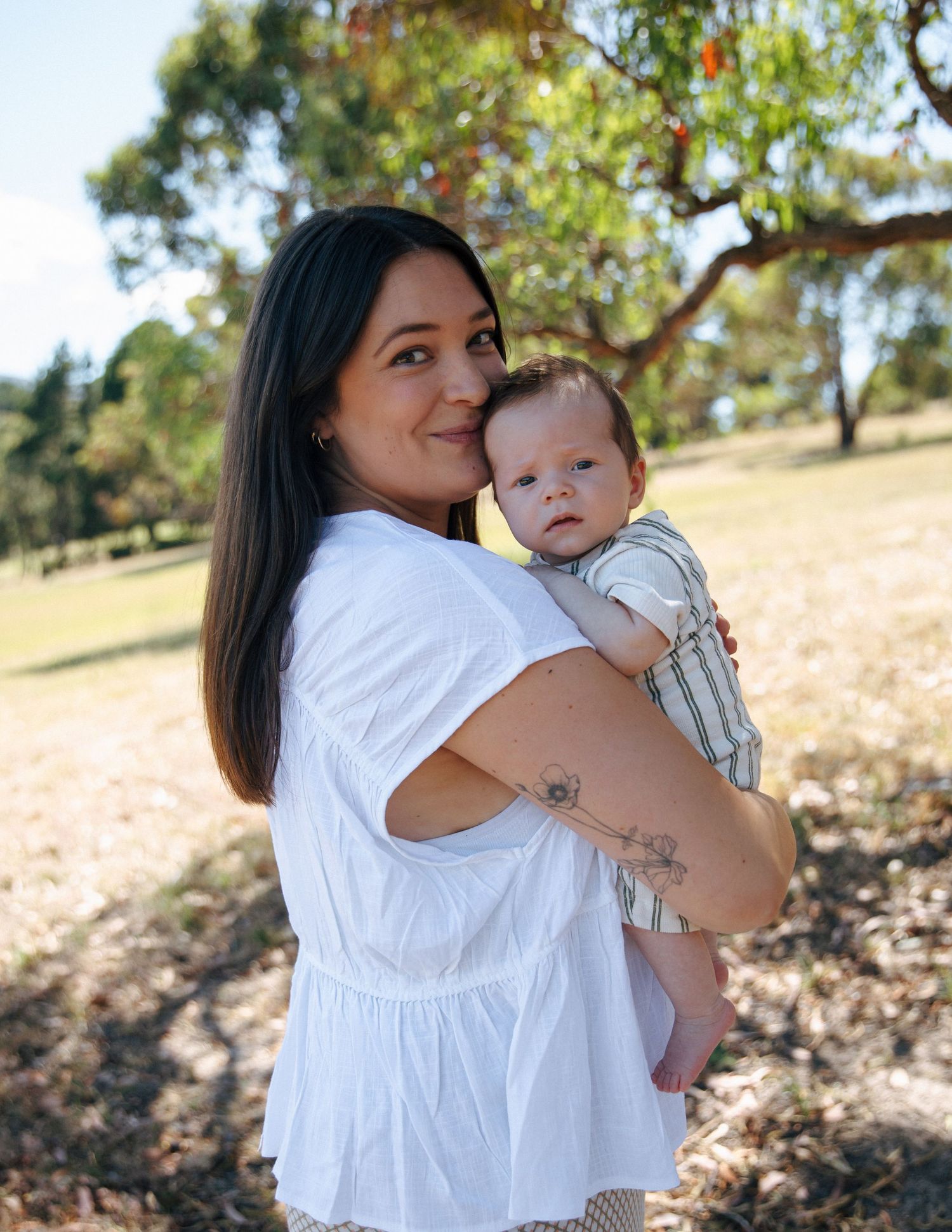 A dark-haired mother smiles over her shoulder while holding her infant baby in an outdoor park setting.