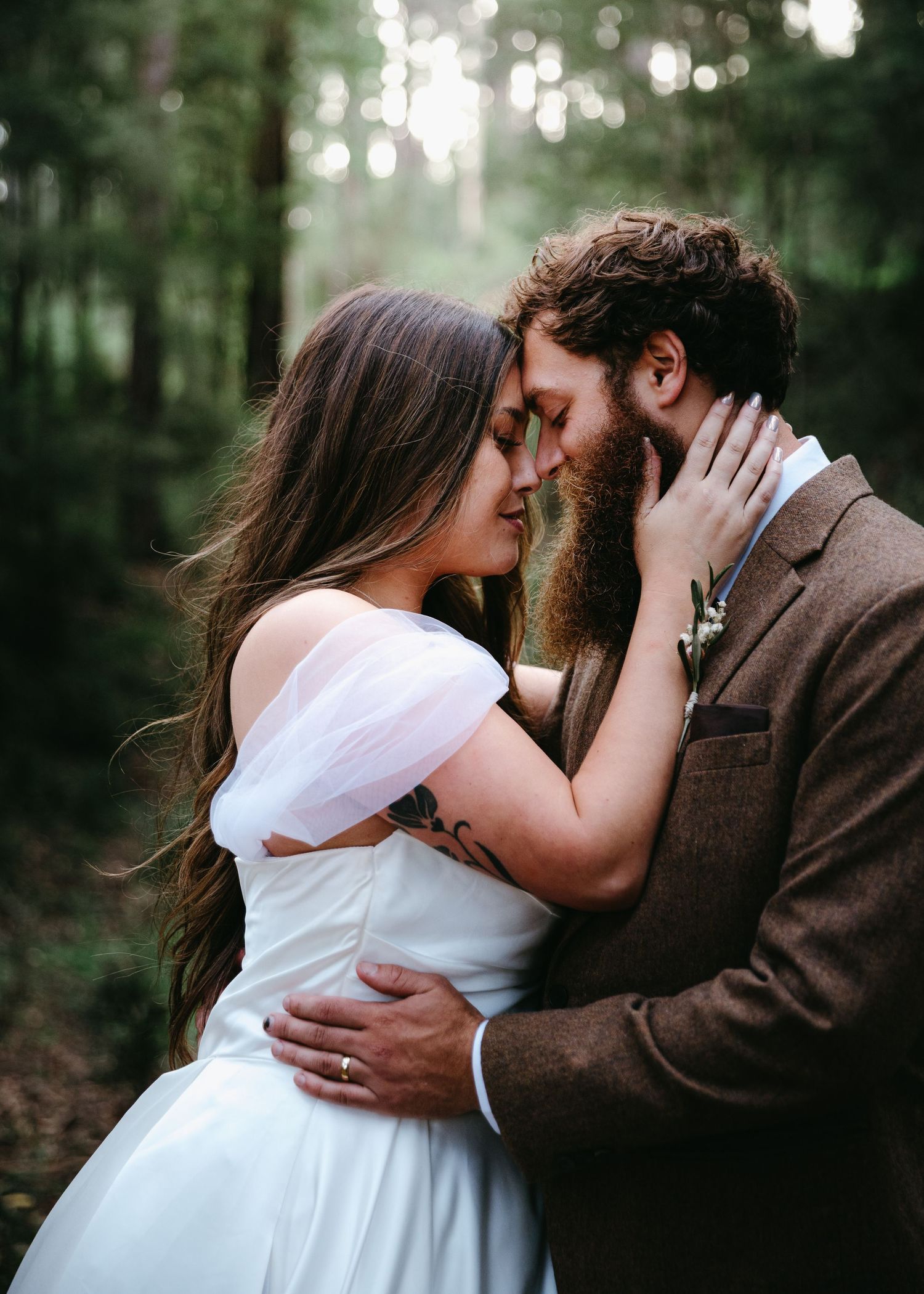 Bride and bearded groom share intimate moment in misty forest setting.