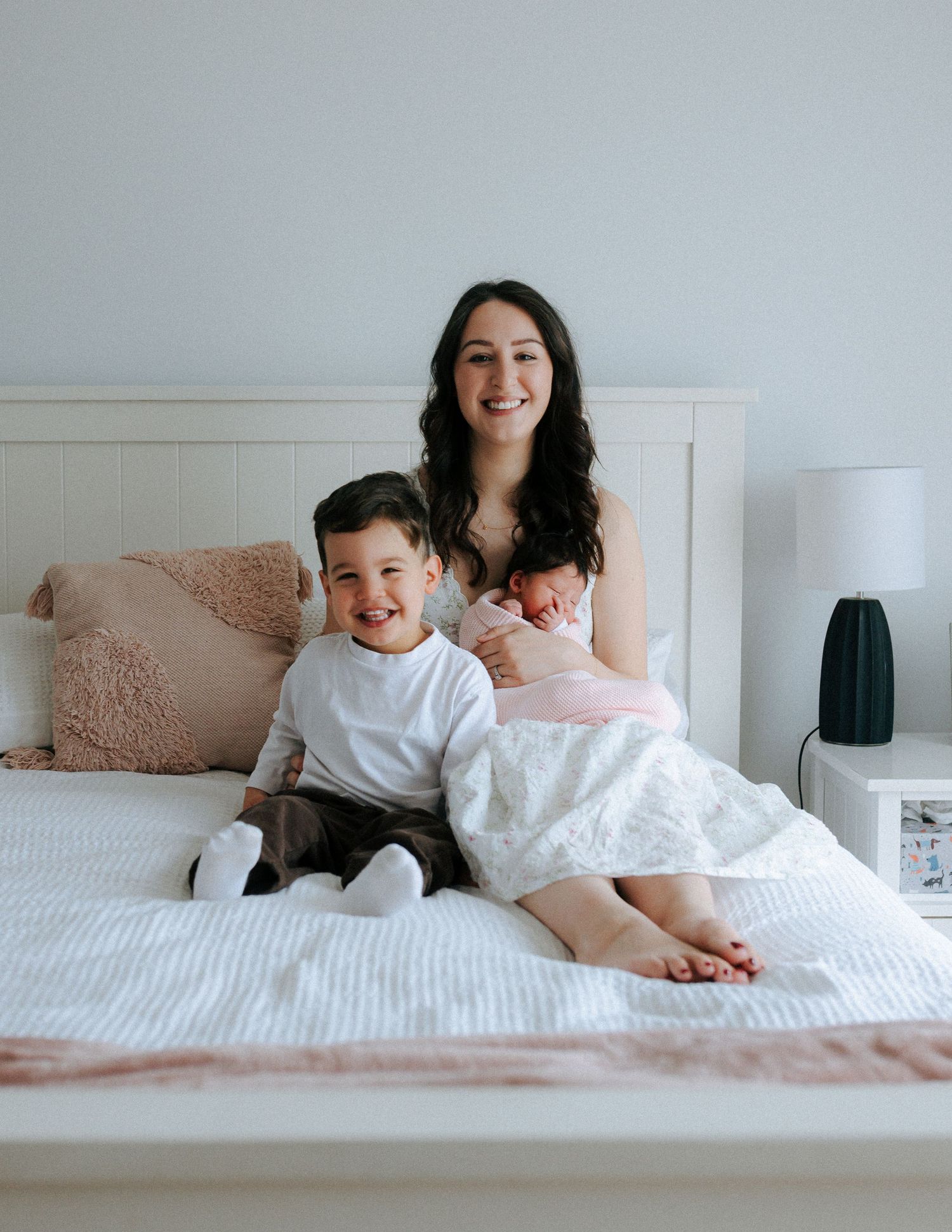 Smiling mother sitting on bed holding newborn baby in pink with toddler son beside her.