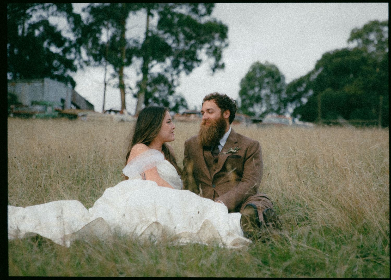 Bride in white off-shoulder gown and bearded groom in brown suit sitting together in a golden wheat field.