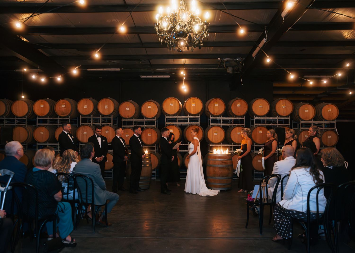 Wedding ceremony inside rustic winery with oak barrel backdrop.