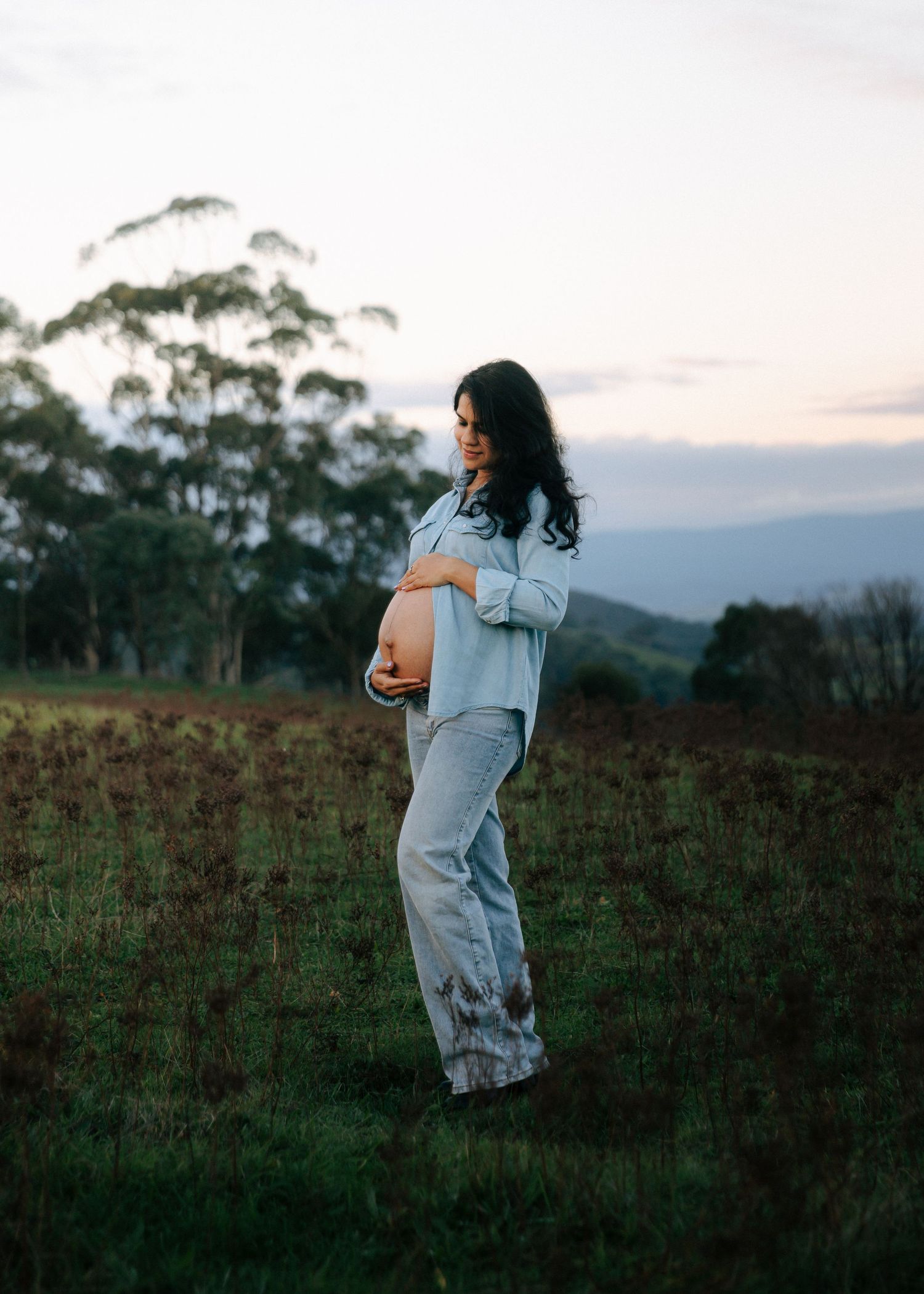 Pregnant woman in light blue denim outfit standing in field at dusk holding baby bump.