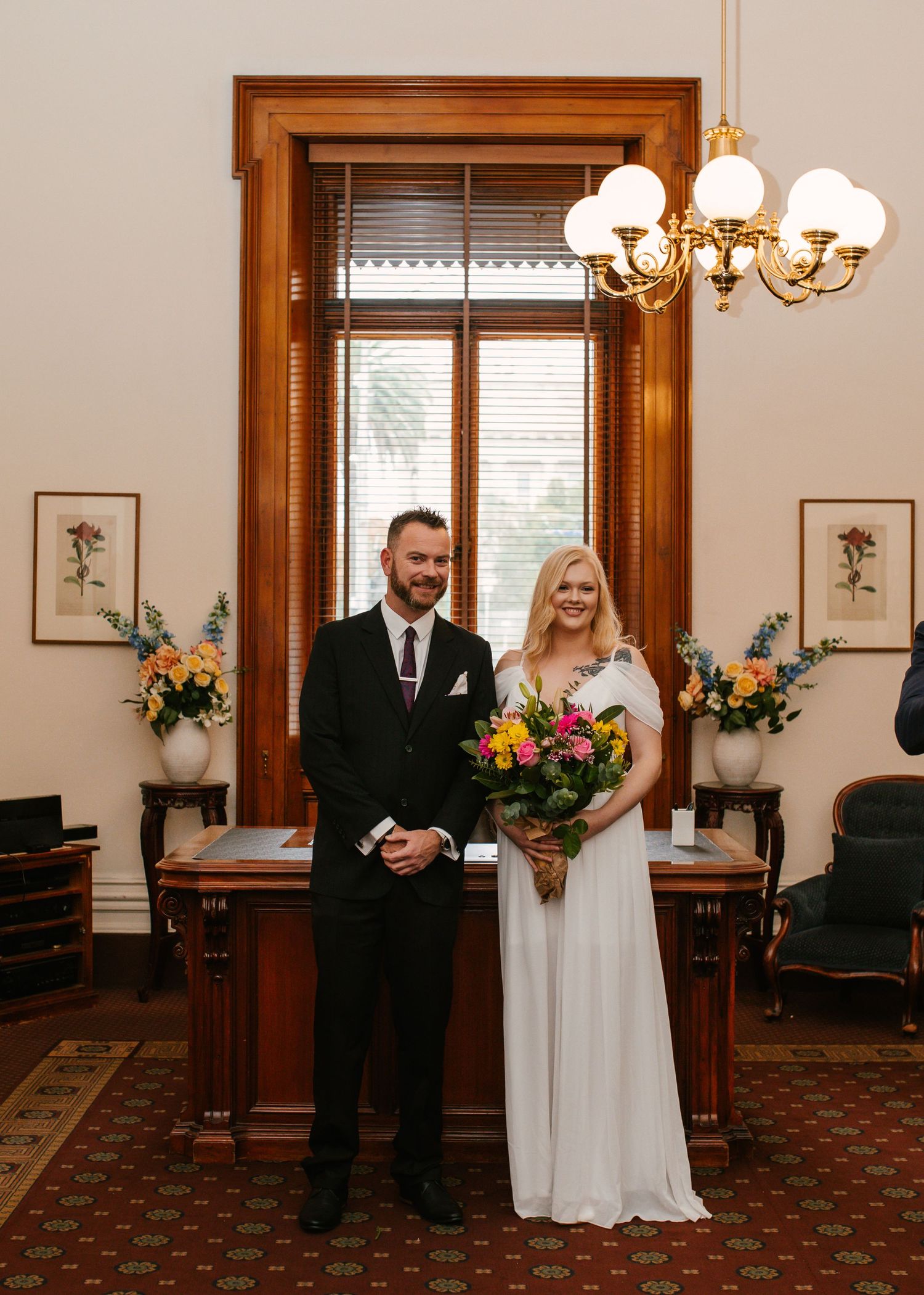 Bride and groom pose in elegant wood-paneled registry office with floral arrangements and chandelier.