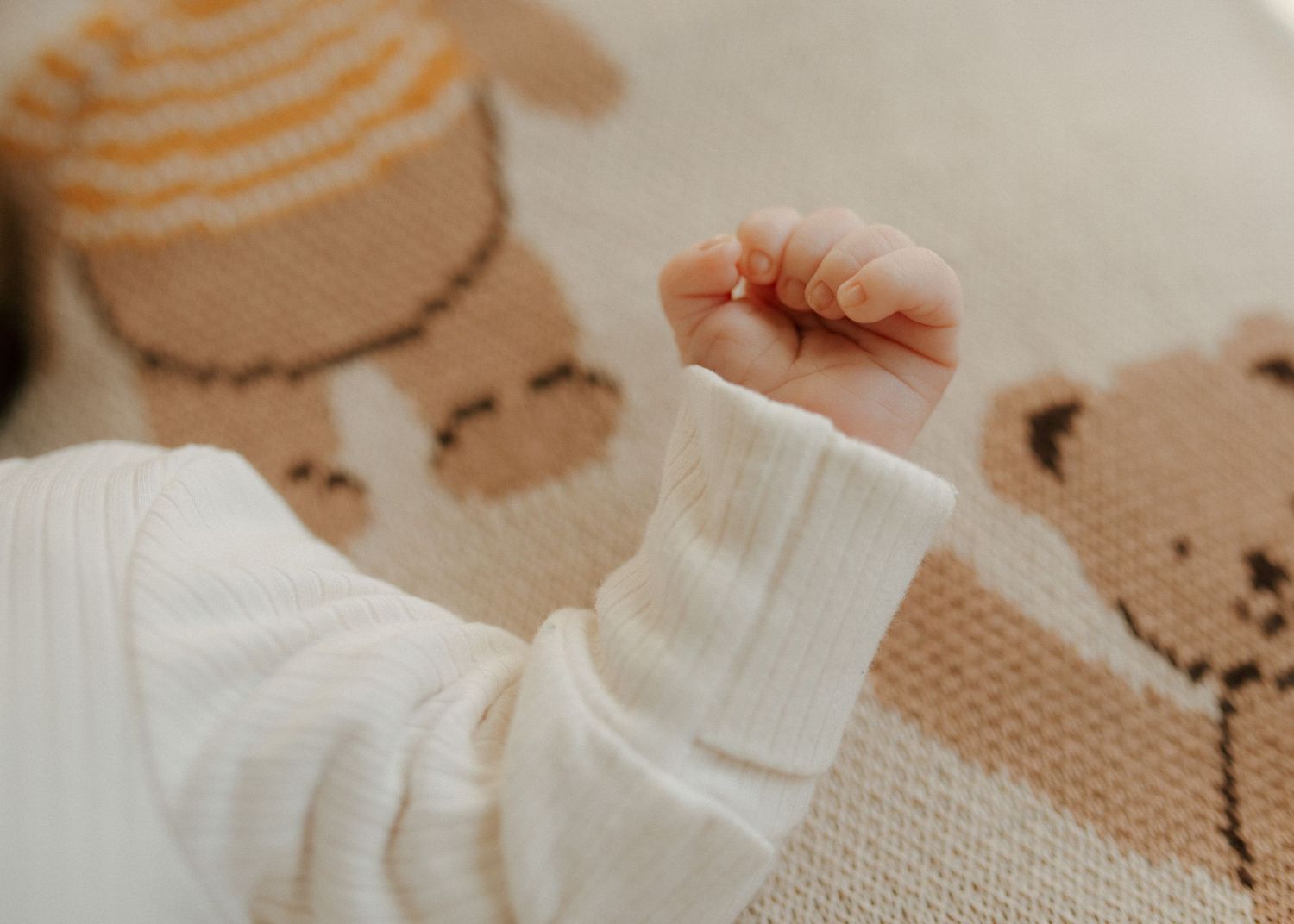 Close-up of a newborn baby's tiny fist in a white outfit on a knitted bear blanket.