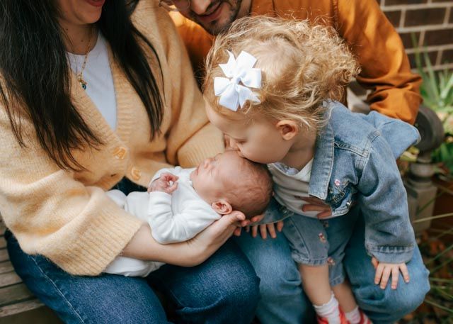 Toddler girl with white bow kissing newborn sibling held by mother outdoors.