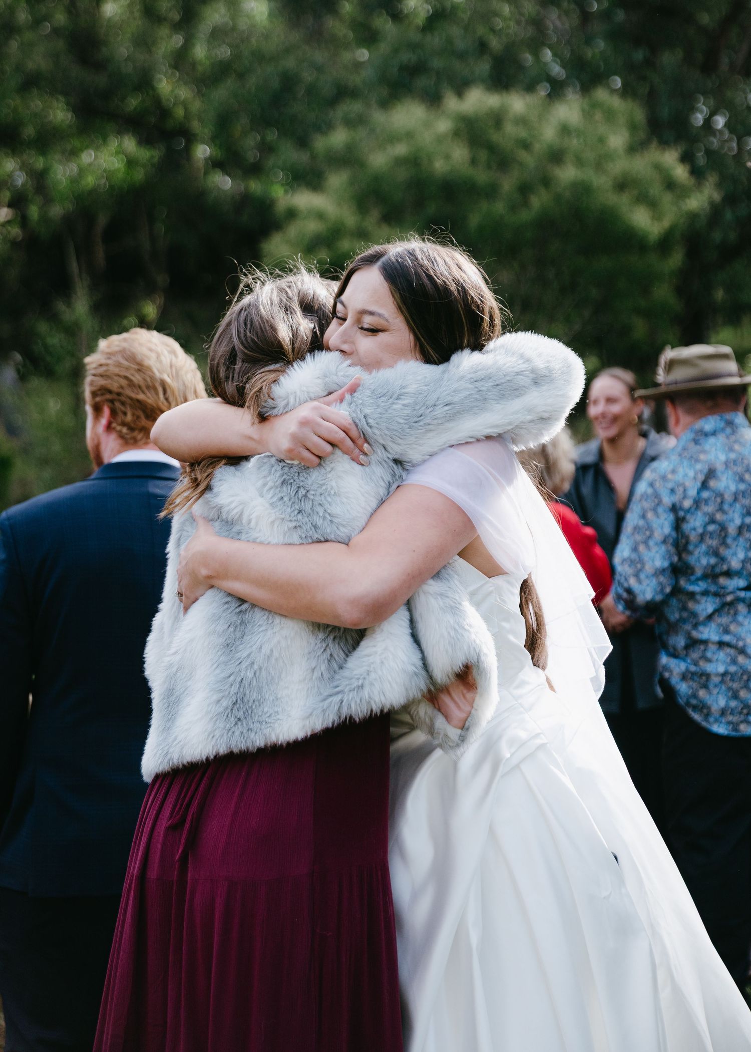 Bride in white gown and fur wrap sharing warm embrace with guest at outdoor wedding celebration.