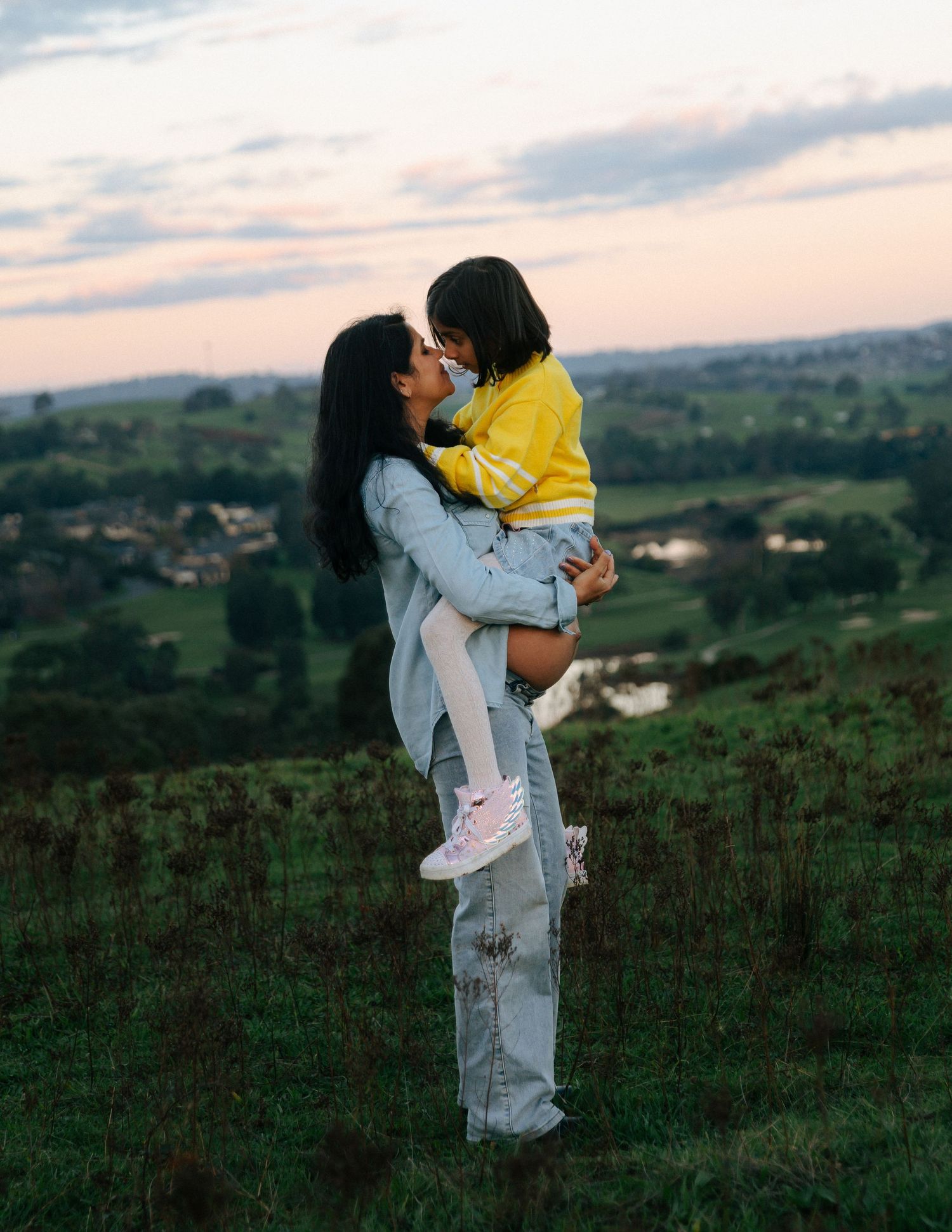 Mother lifting and kissing her young child in a yellow jacket on a scenic green hillside at dusk.