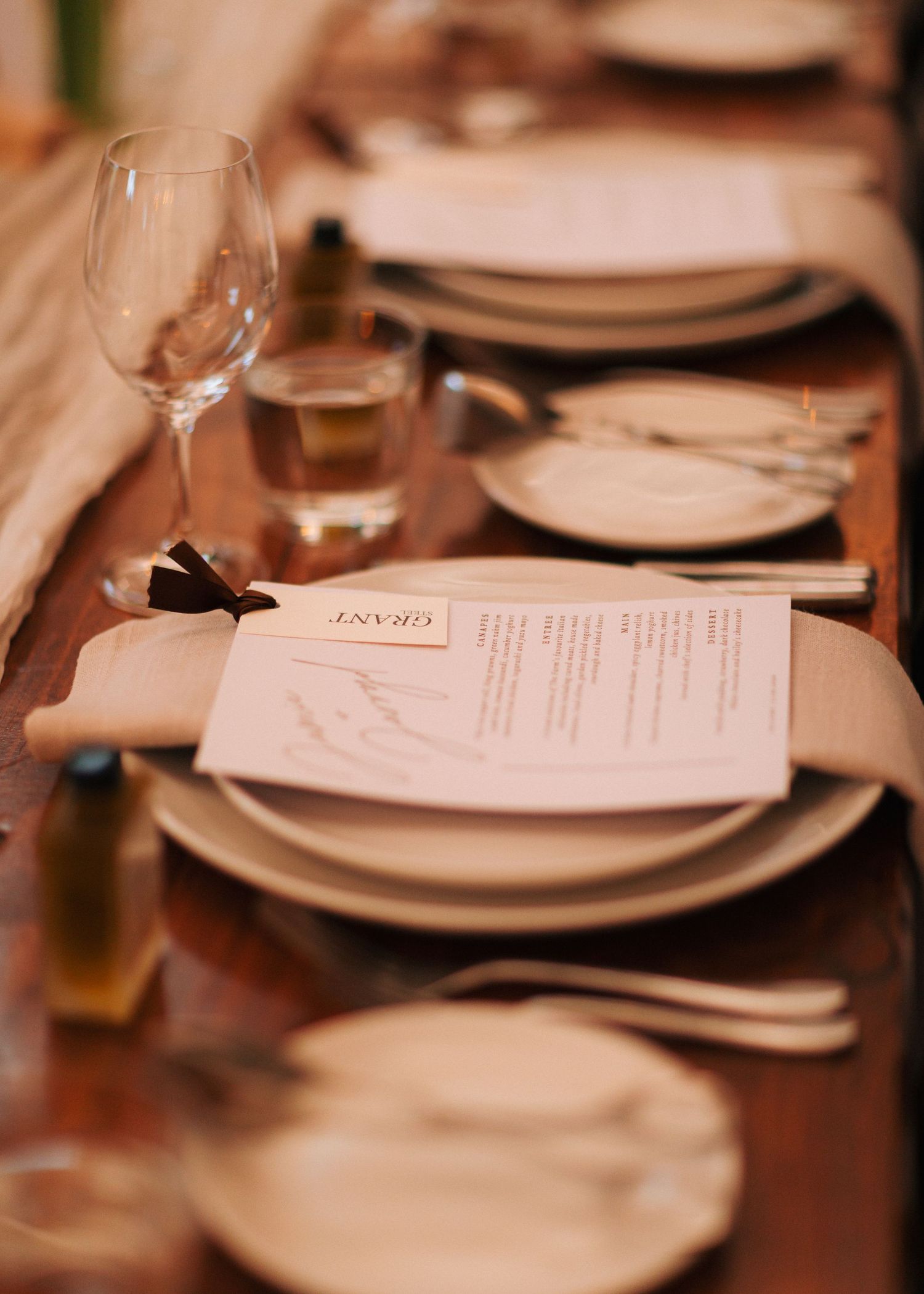 Rustic wedding place setting with stacked plates, personalized menu card, linen napkin, and glassware on wooden table.