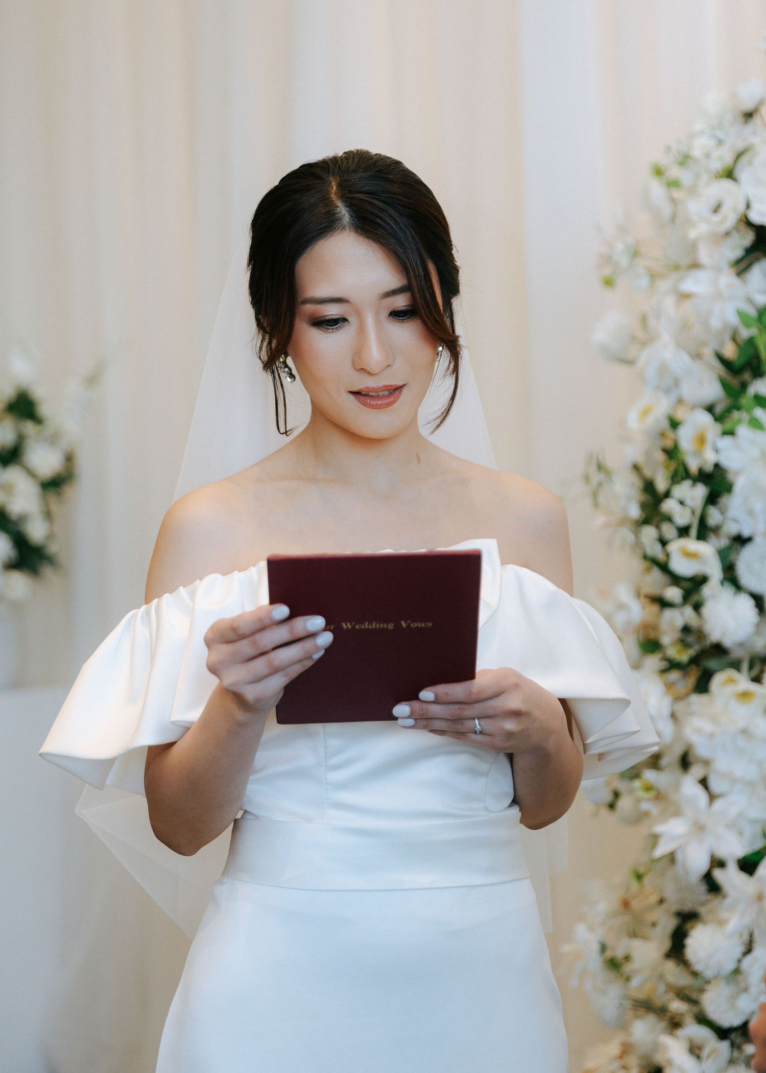Beautiful bride in off-shoulder gown reading wedding vows from a burgundy booklet at the altar.