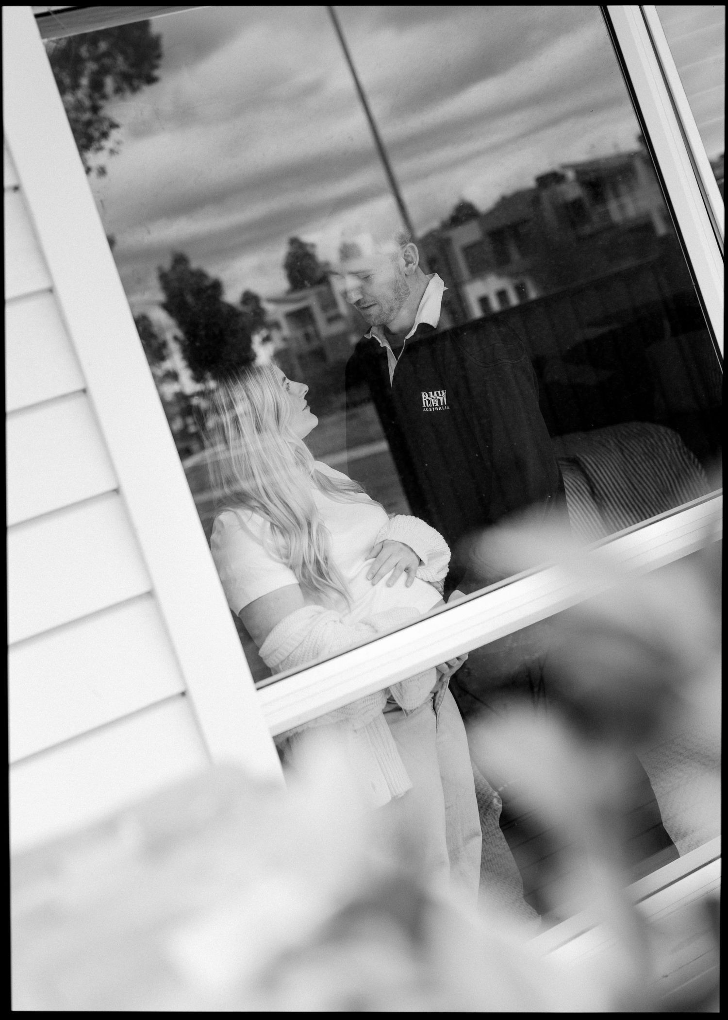 Black and white photo of couple sharing intimate moment captured through a reflective window pane.