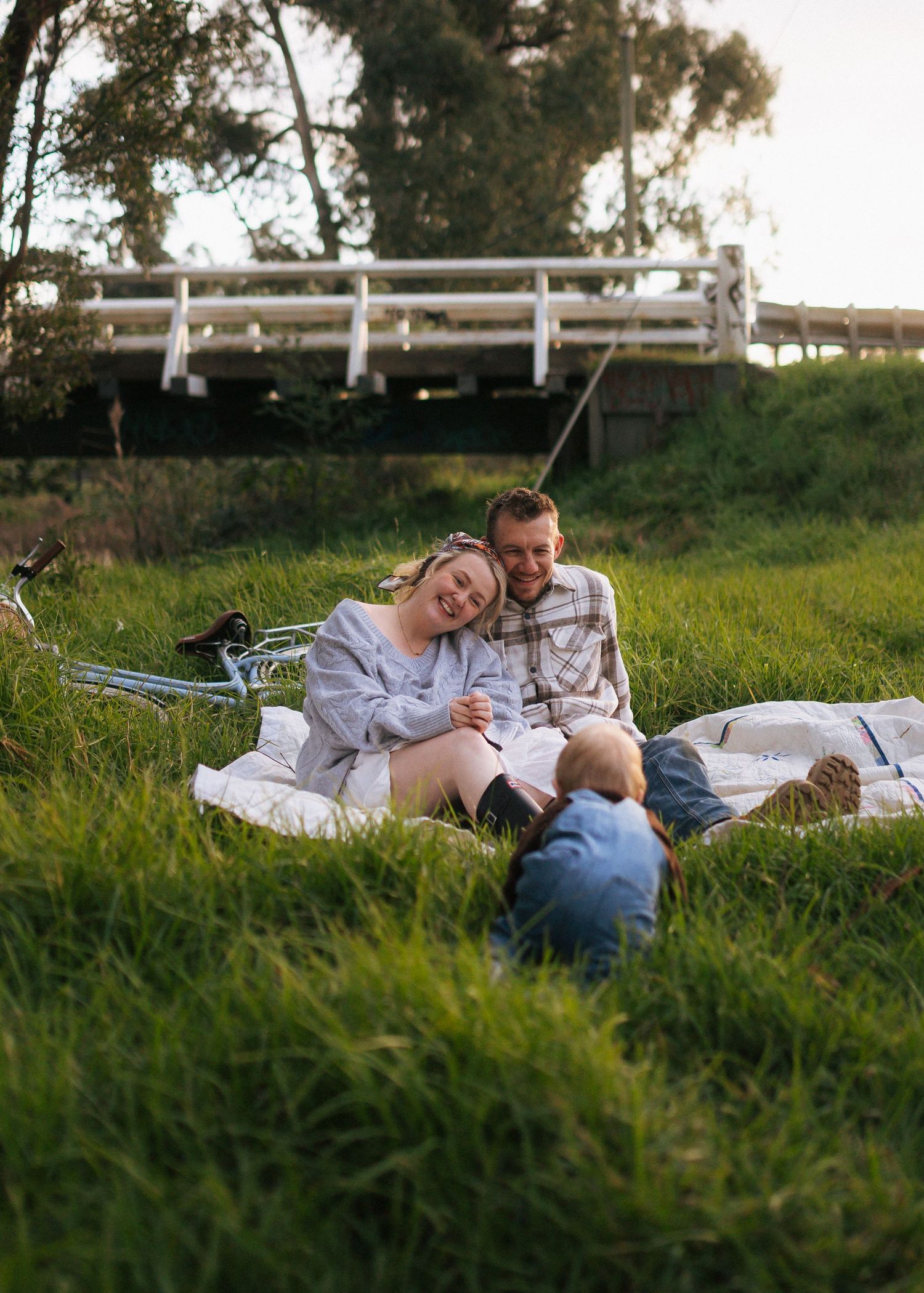 Couple relaxing on a blanket in lush green grass while toddler plays nearby with a bridge backdrop.