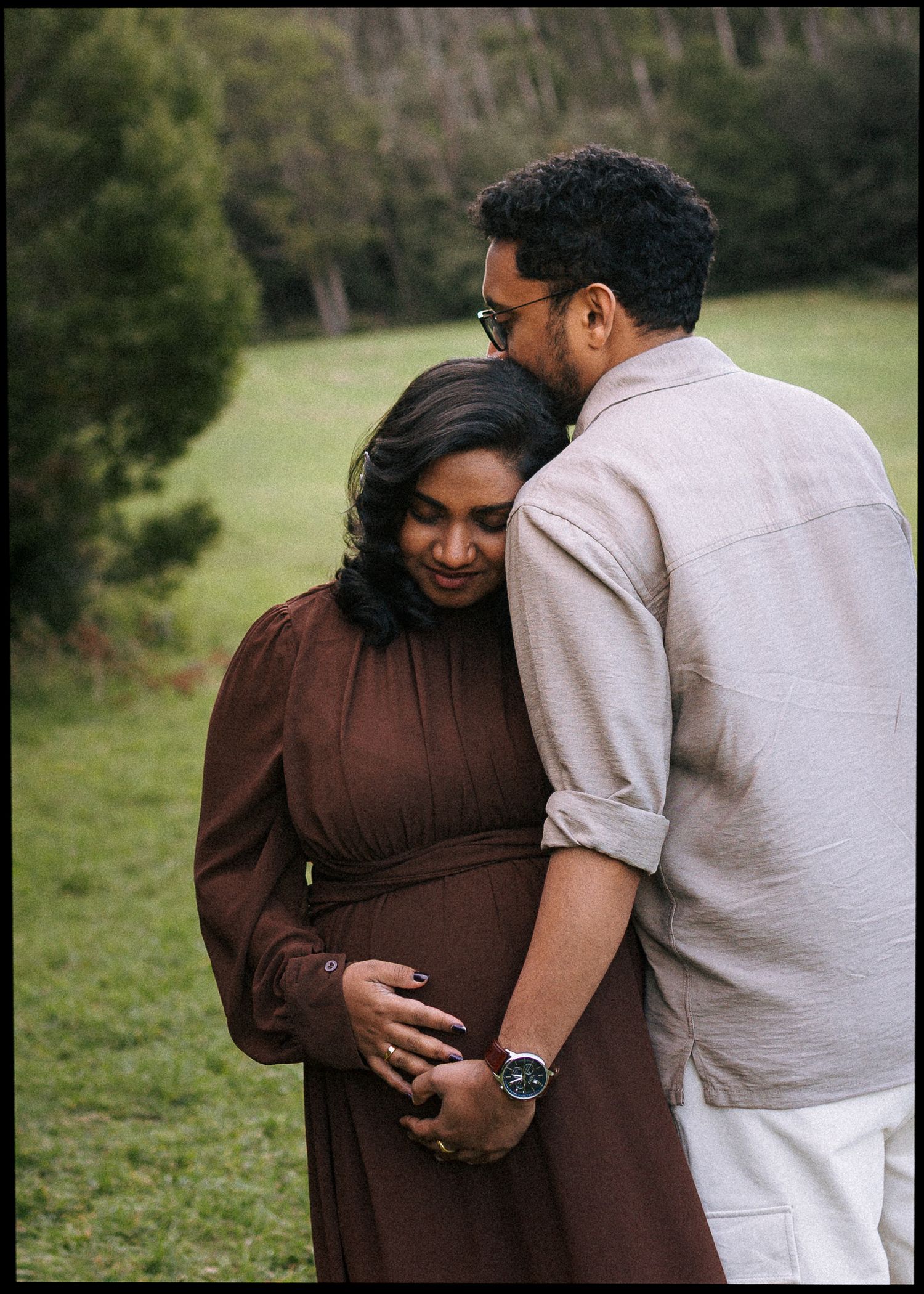 Expecting couple embracing in a green field, the woman in a brown dress cradling her baby bump as the man in a gray shirt hugs her.