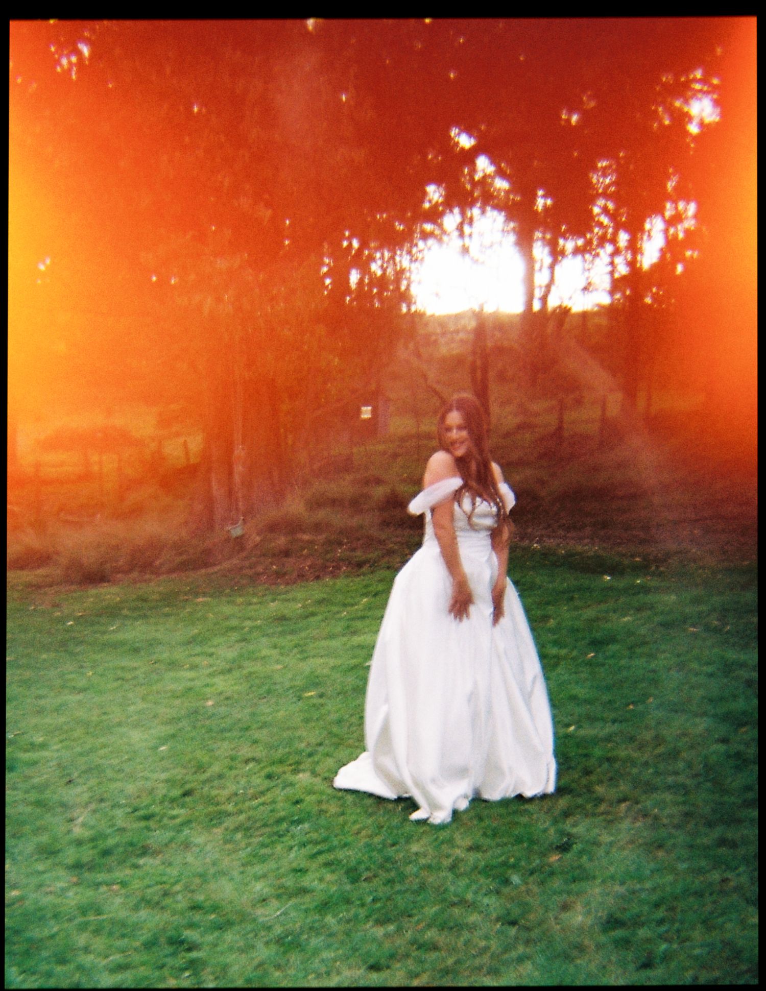 Bride in an off-shoulder white ball gown posing at golden sunset with dramatic orange lens flare in garden.