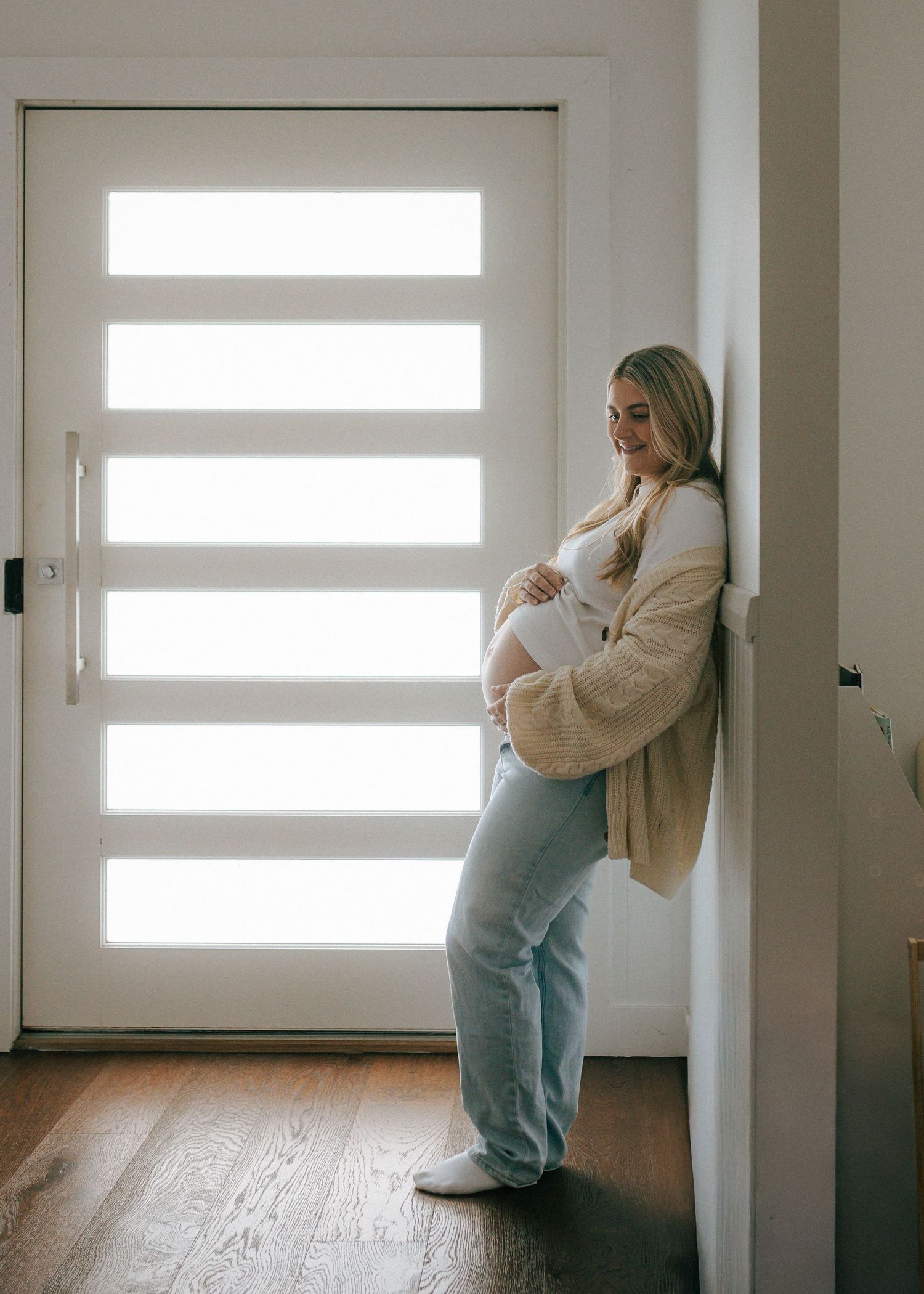 Pregnant woman in cream cardigan leaning against wall by front door cradling baby bump.