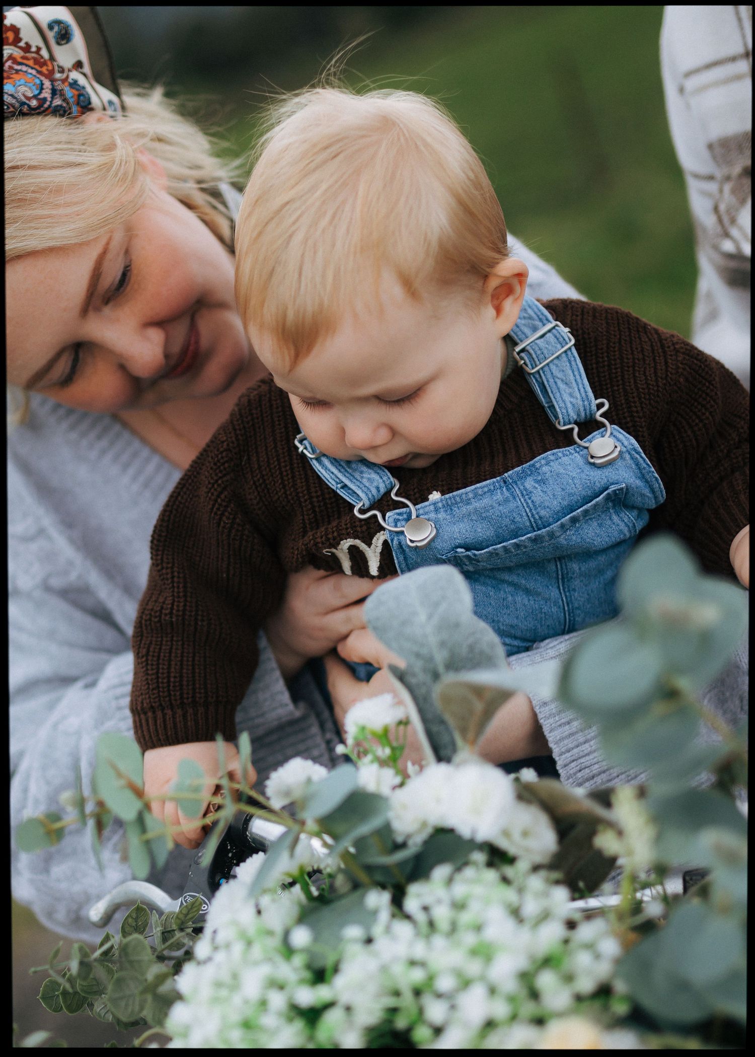 Loving mother and red-haired toddler in denim overalls exploring white flowers on a bicycle.