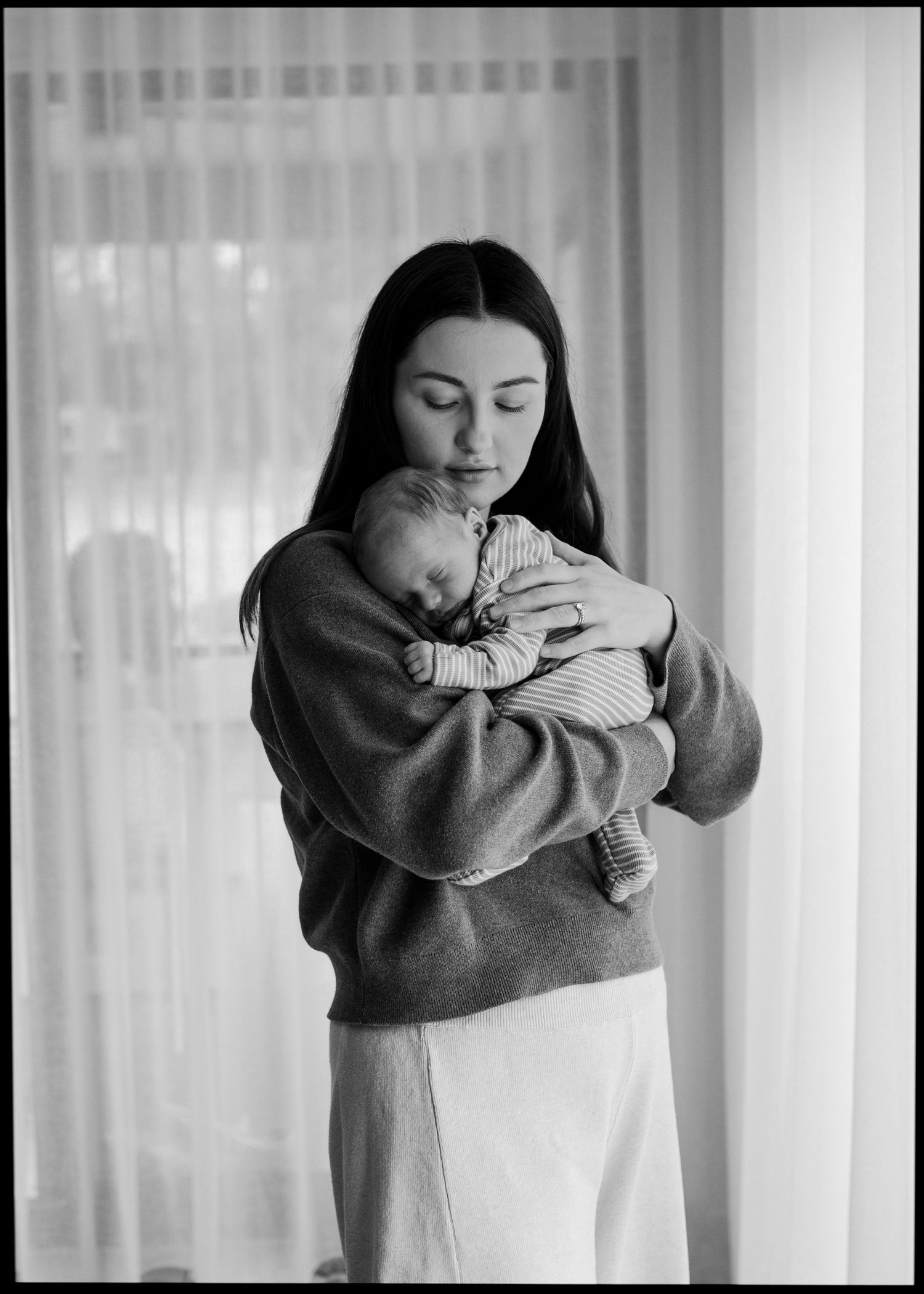 Black and white portrait of a mother in a grey sweater cradling her sleeping newborn baby.