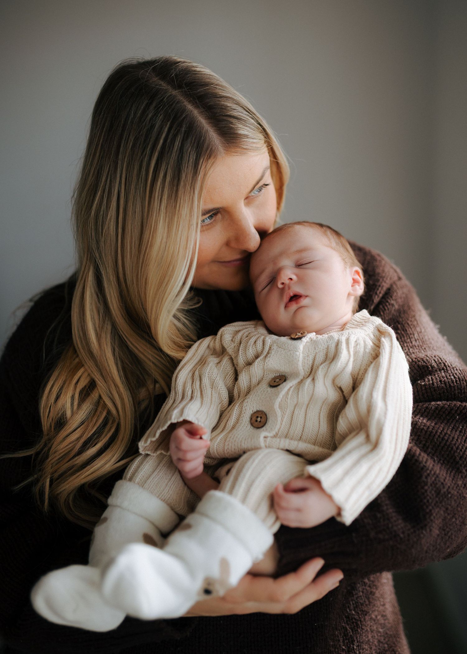 Mother in brown sweater gently kissing sleeping newborn dressed in cream cable-knit cardigan and white booties.