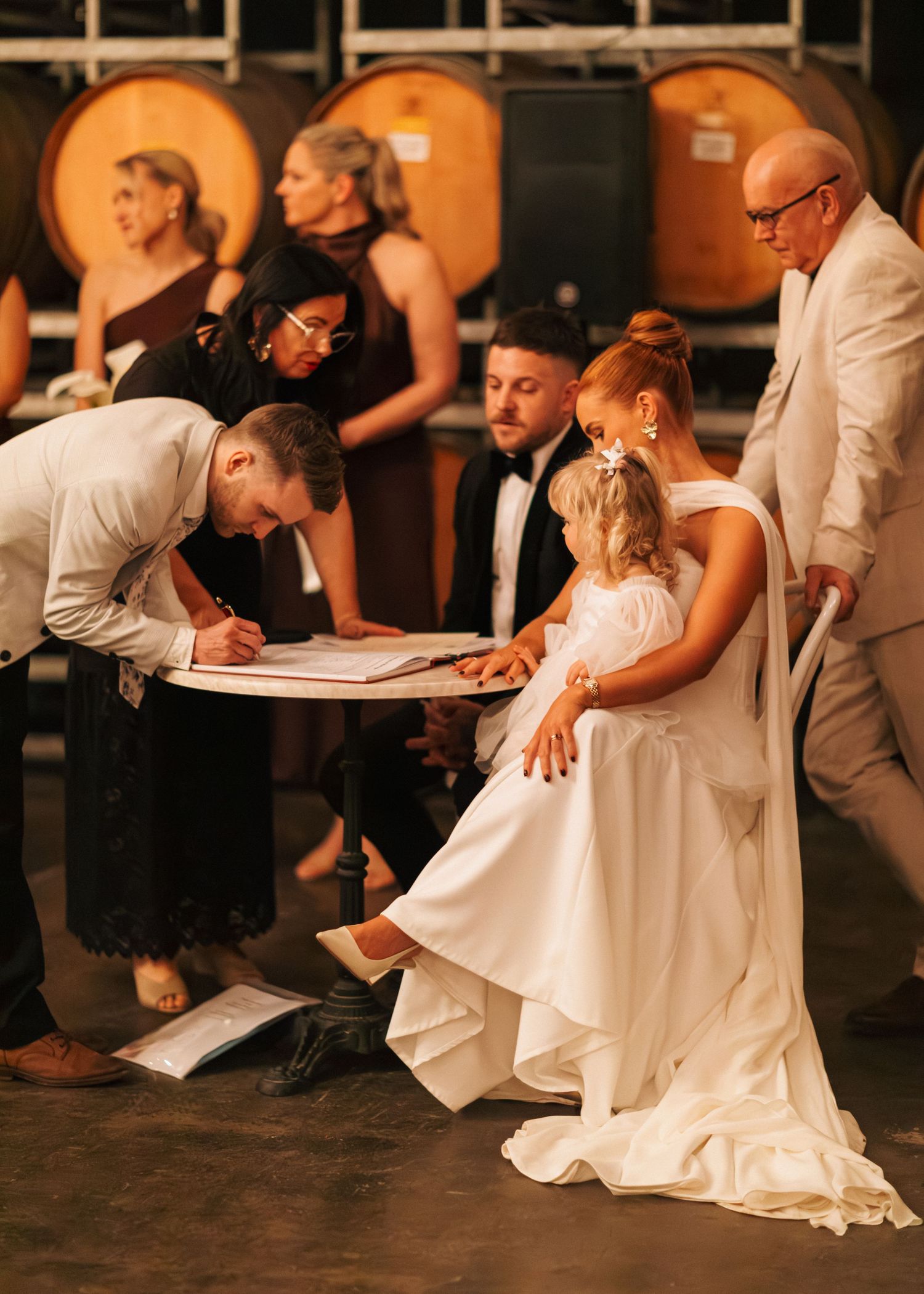 Bride and groom signing wedding registry at a winery venue with guests and flower girl watching.