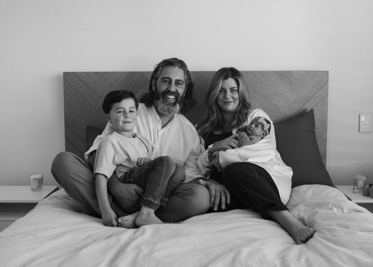 Black and white family portrait of parents with toddler son and newborn baby on a bed.
