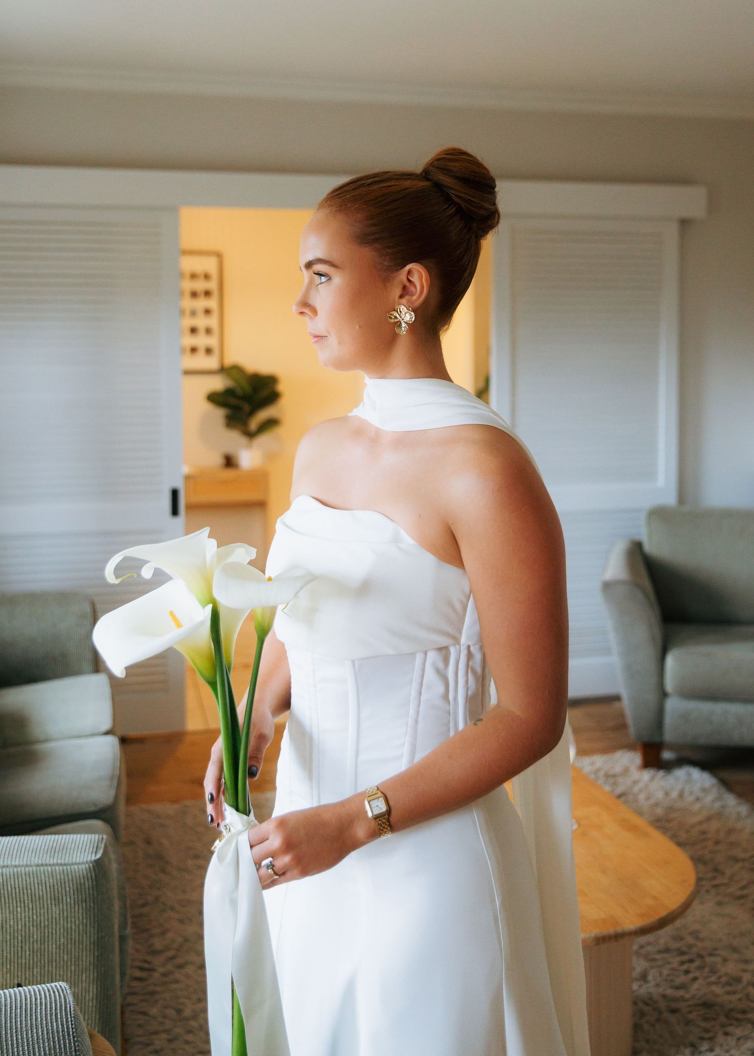 Bride in white strapless corset gown holds calla lily bouquet while gazing out window.