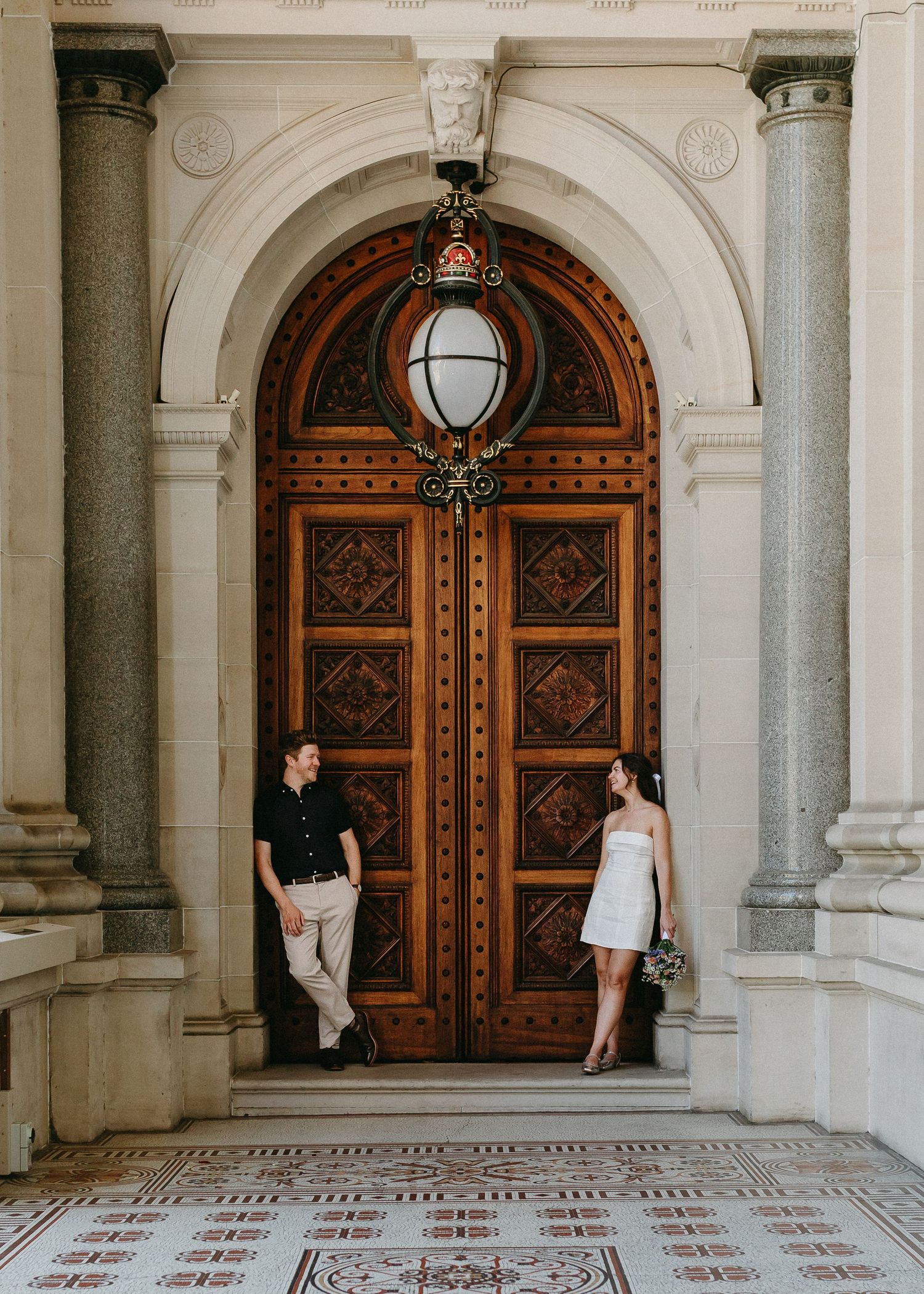 Bride and groom pose beside ornate wooden arched door of elegant heritage building.