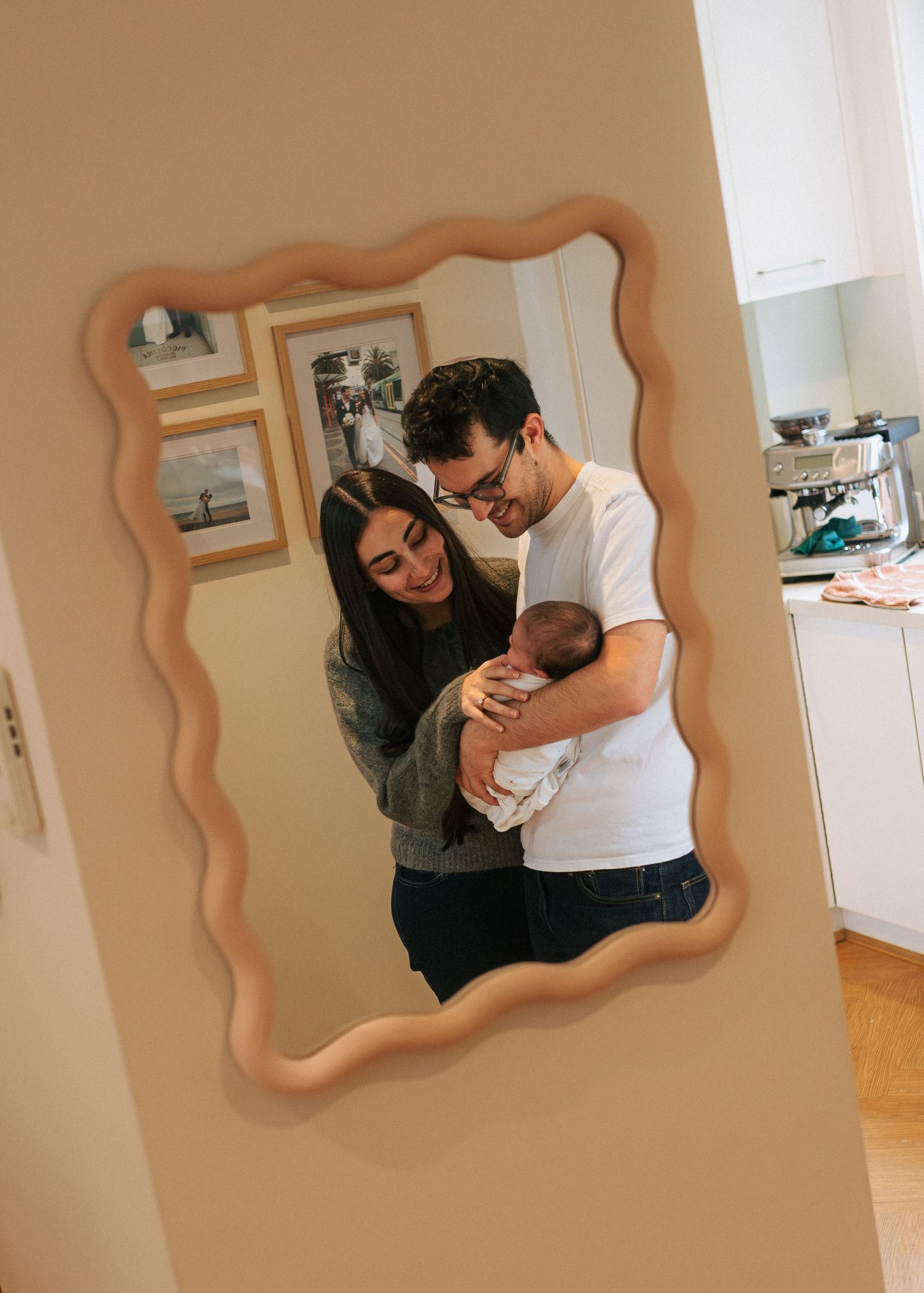 New parents smiling and holding their newborn baby reflected in a decorative mirror.
