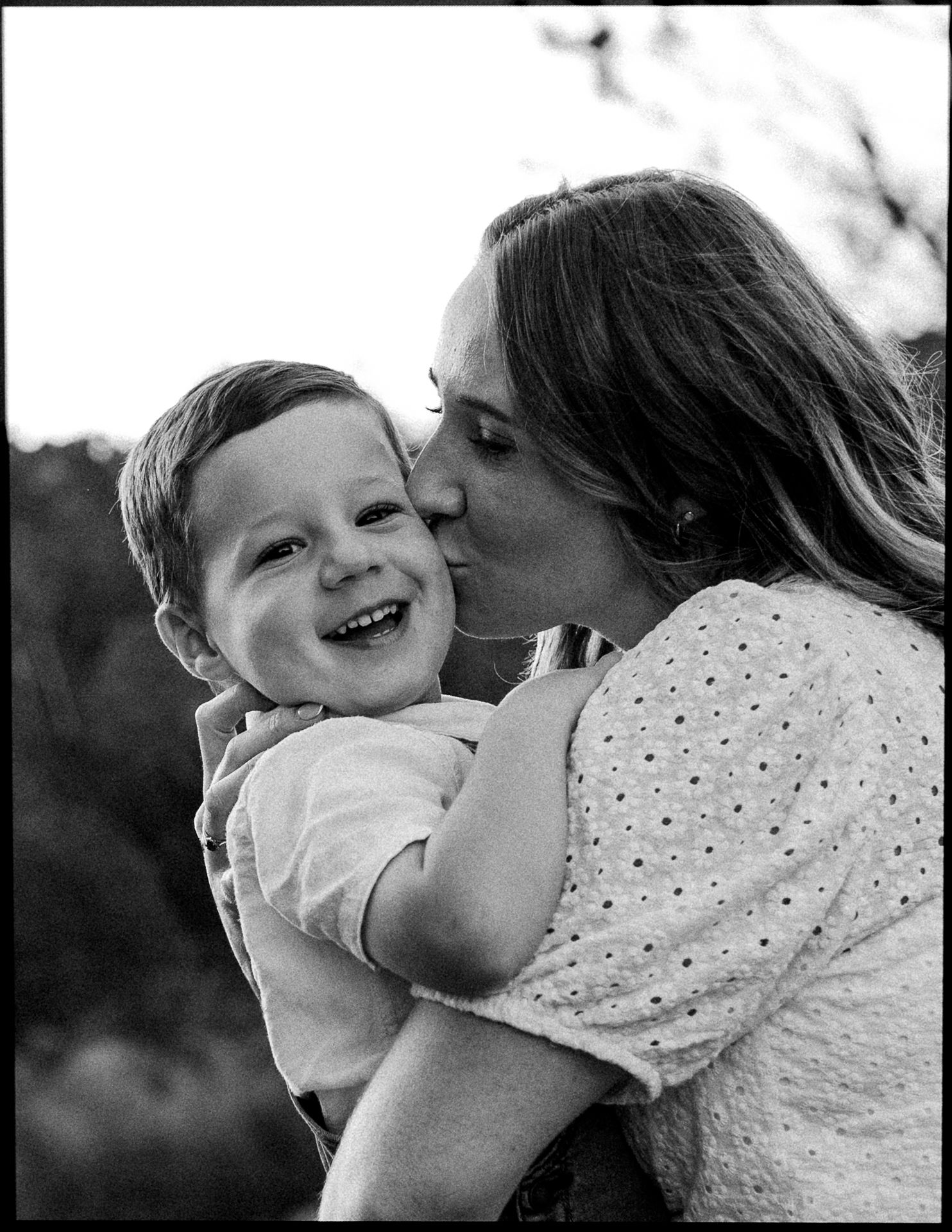 Black and white photo of a mother kissing her smiling young son outdoors.