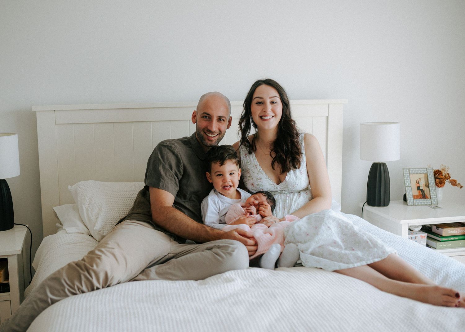 Happy family portrait on bed with parents smiling, toddler and newborn baby in pink outfit.